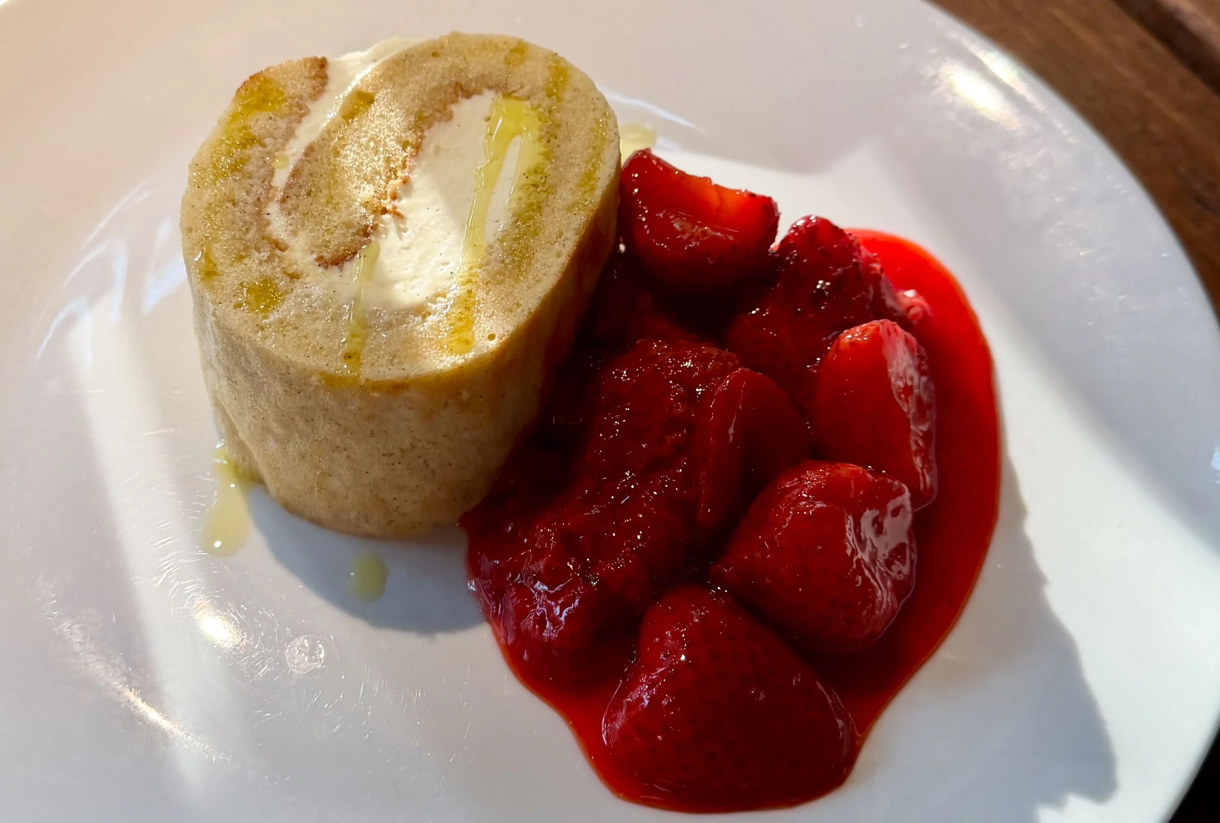 A dessert with a rolled sponge cake filled with cream, topped with a drizzle of honey or syrup, served with a side of fresh strawberries and strawberry sauce on a white plate.