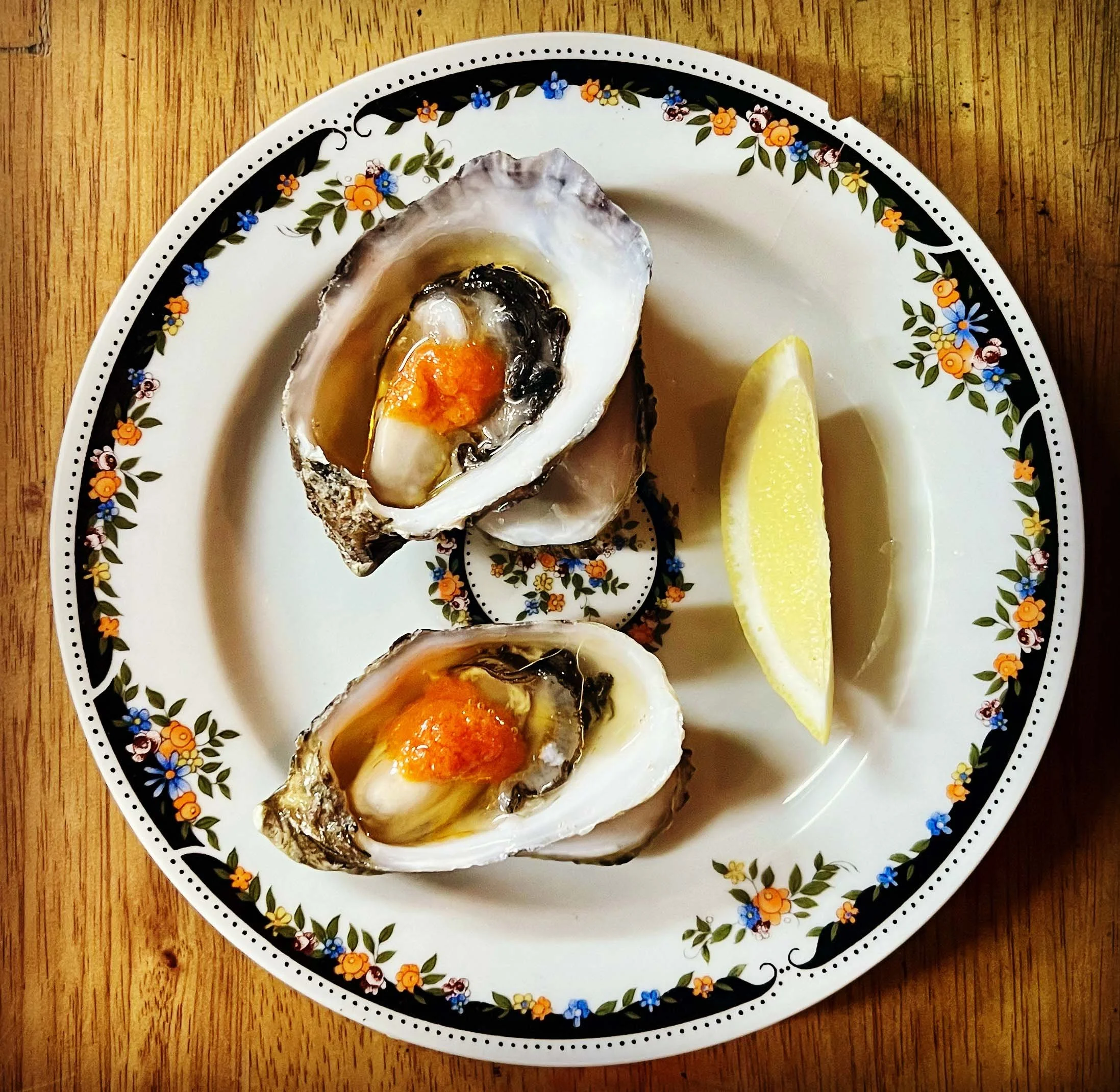 A plate with three oysters, lemon wedges, and a sauce or topping on the oysters, on a wooden surface.