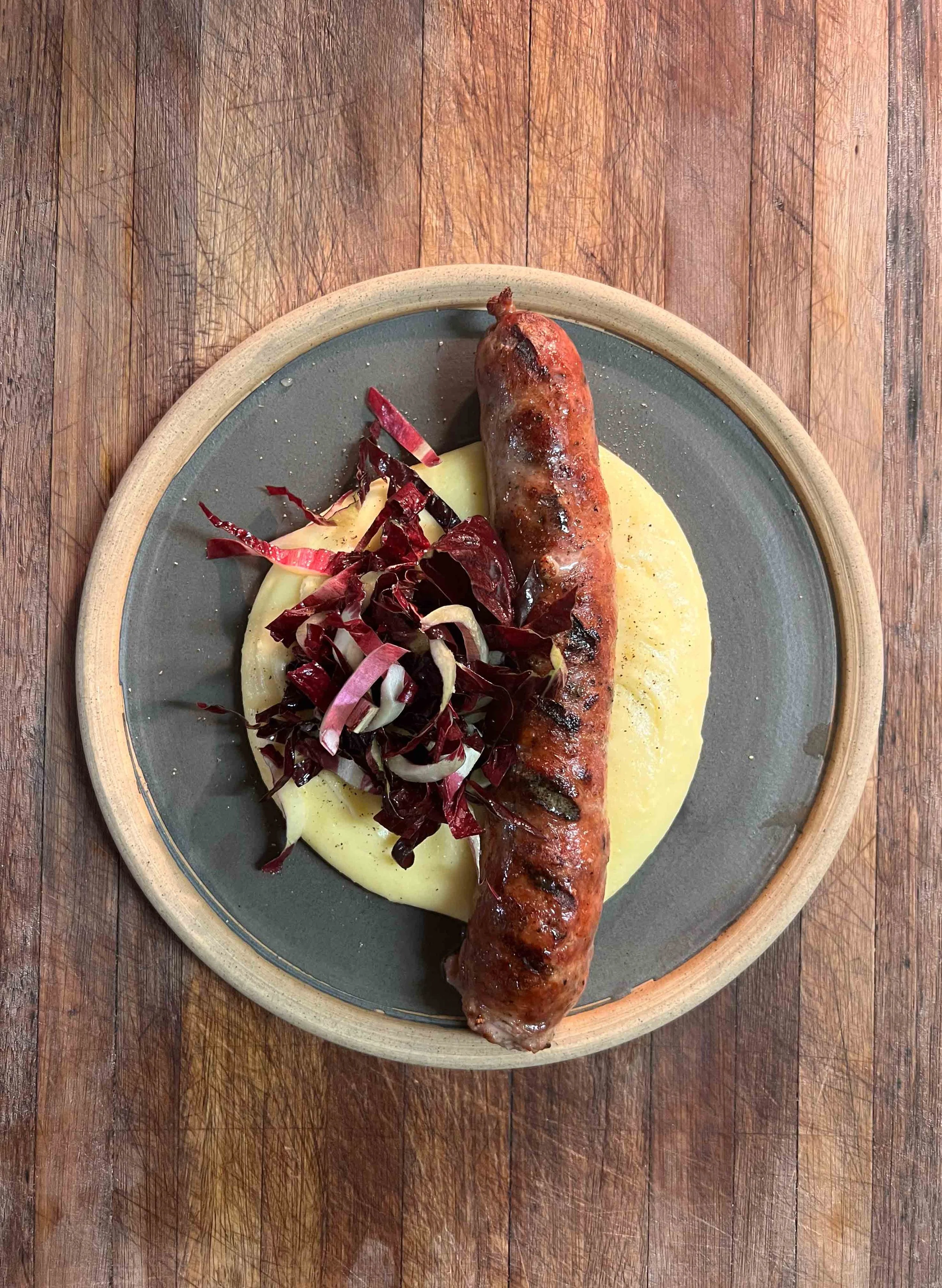 A plate with a grilled sausage, mashed potatoes, and a side of radicchio salad on a wooden table.