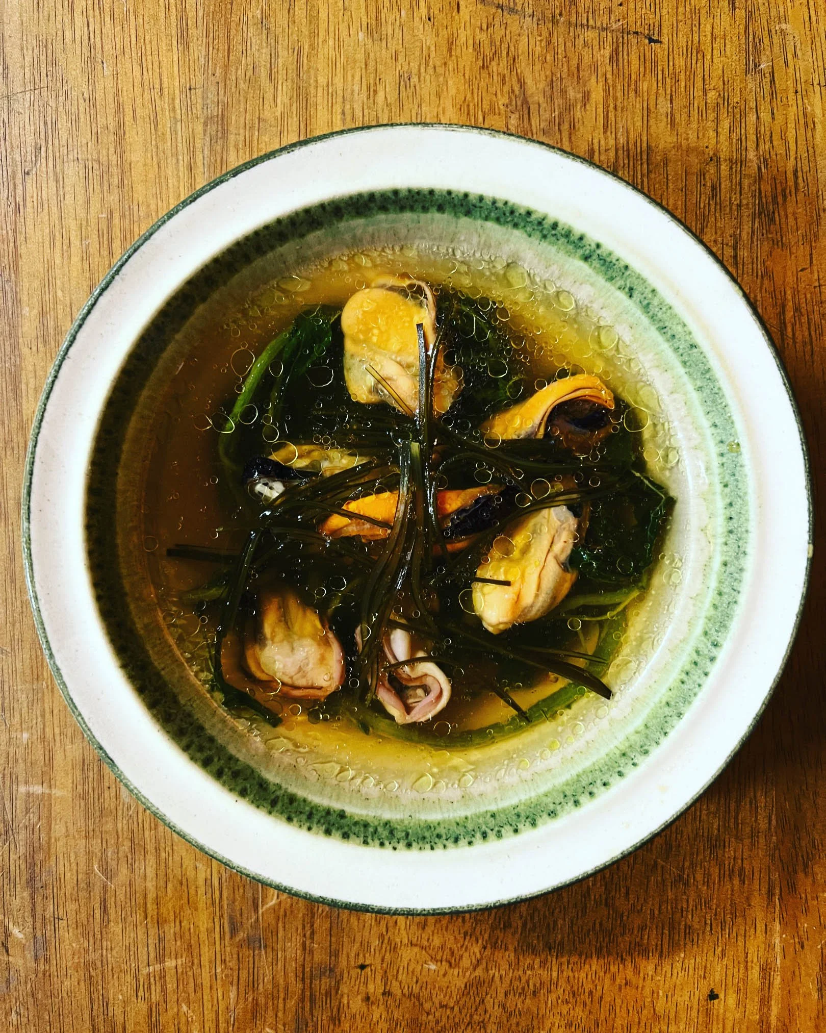 A bowl of clear broth soup with mussels, green herbs, and oil bubbles, placed on a wooden table.