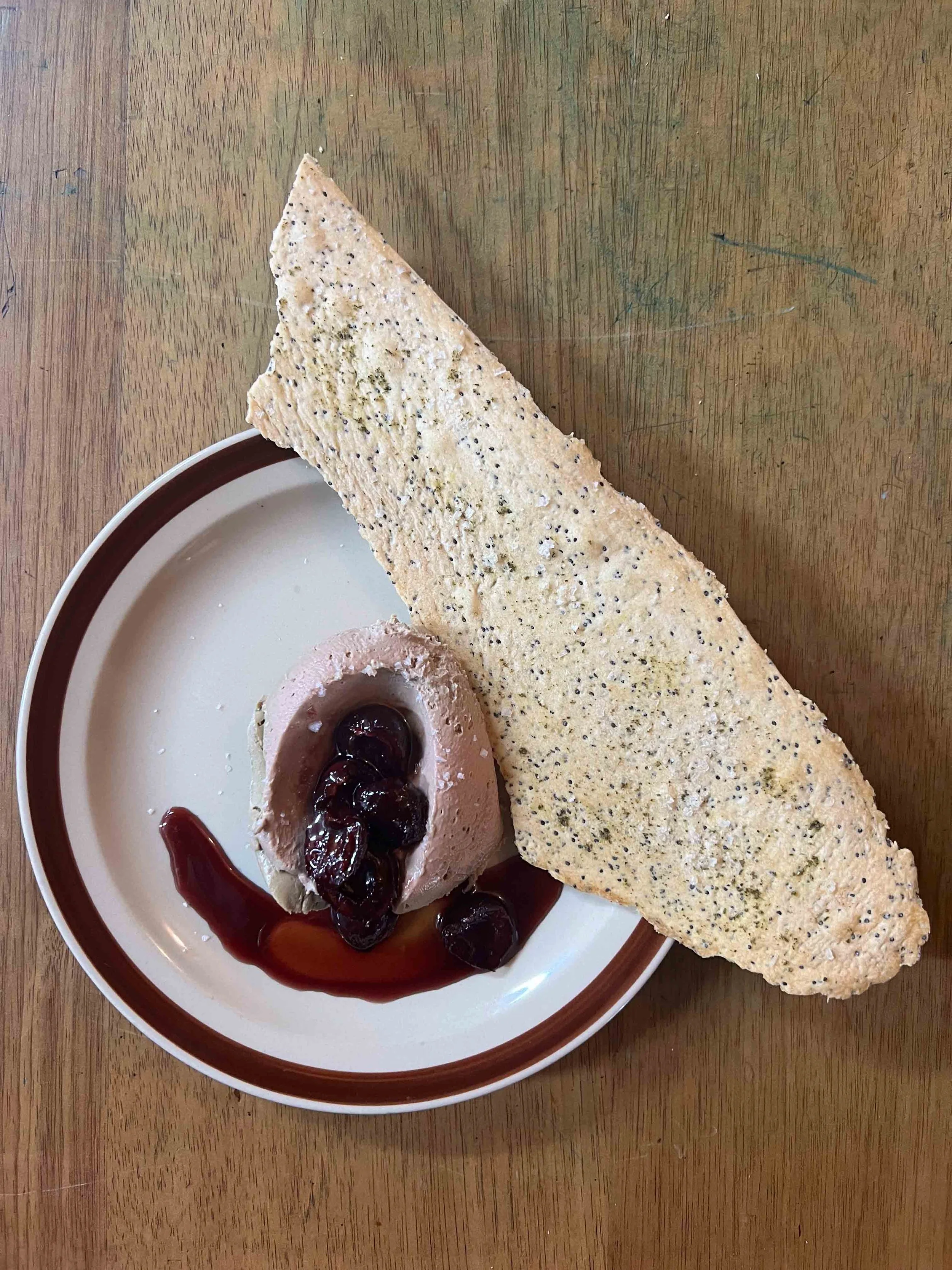 A plate with a large triangular piece of bread, a scoop of pink ice cream topped with dark cherries and syrup, on a wooden table.