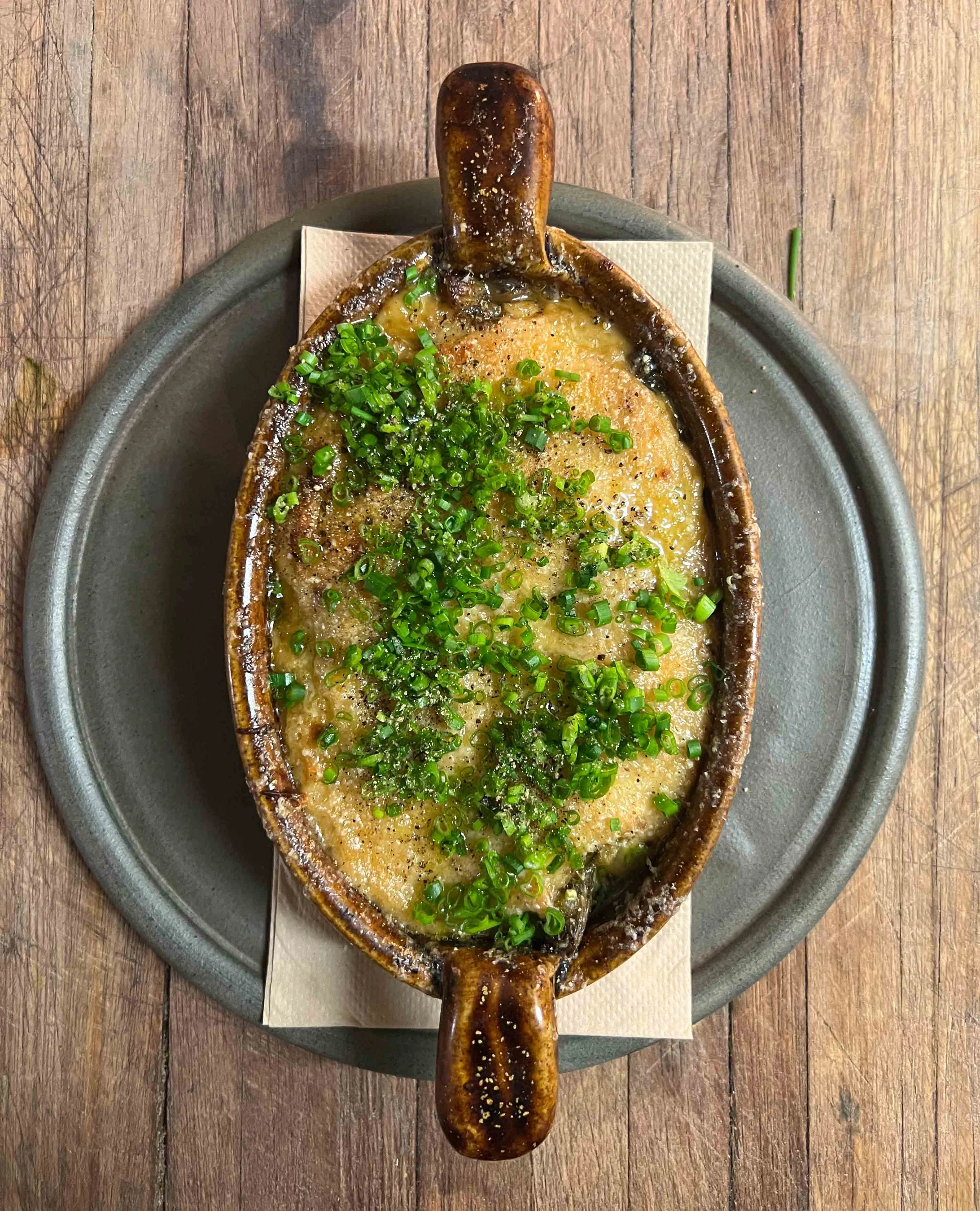A baked potato served in a crispy skin on a round metal plate with chopped green onions and seasoning, placed on a wooden table.