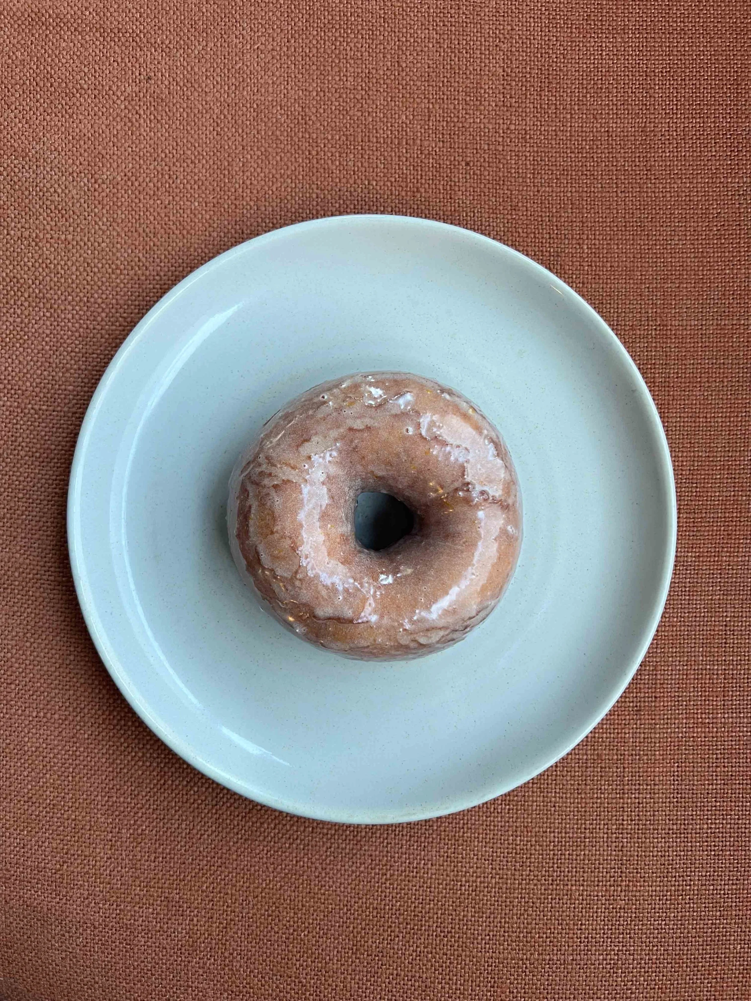 A glazed donut on a white plate placed on a brown textured surface.