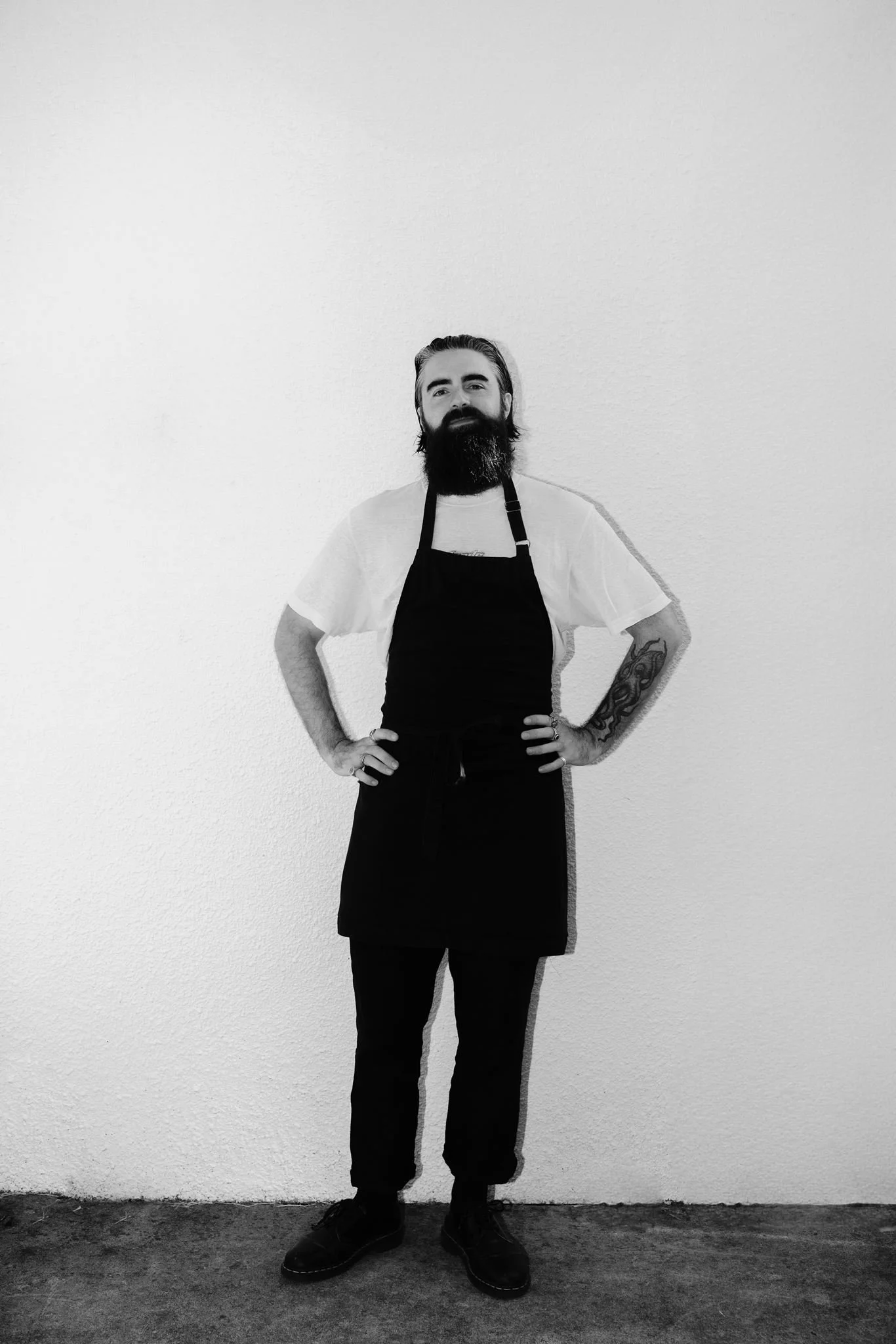 A man with long hair and a beard, wearing a white t-shirt and a black apron, standing with hands on hips against a plain white wall.