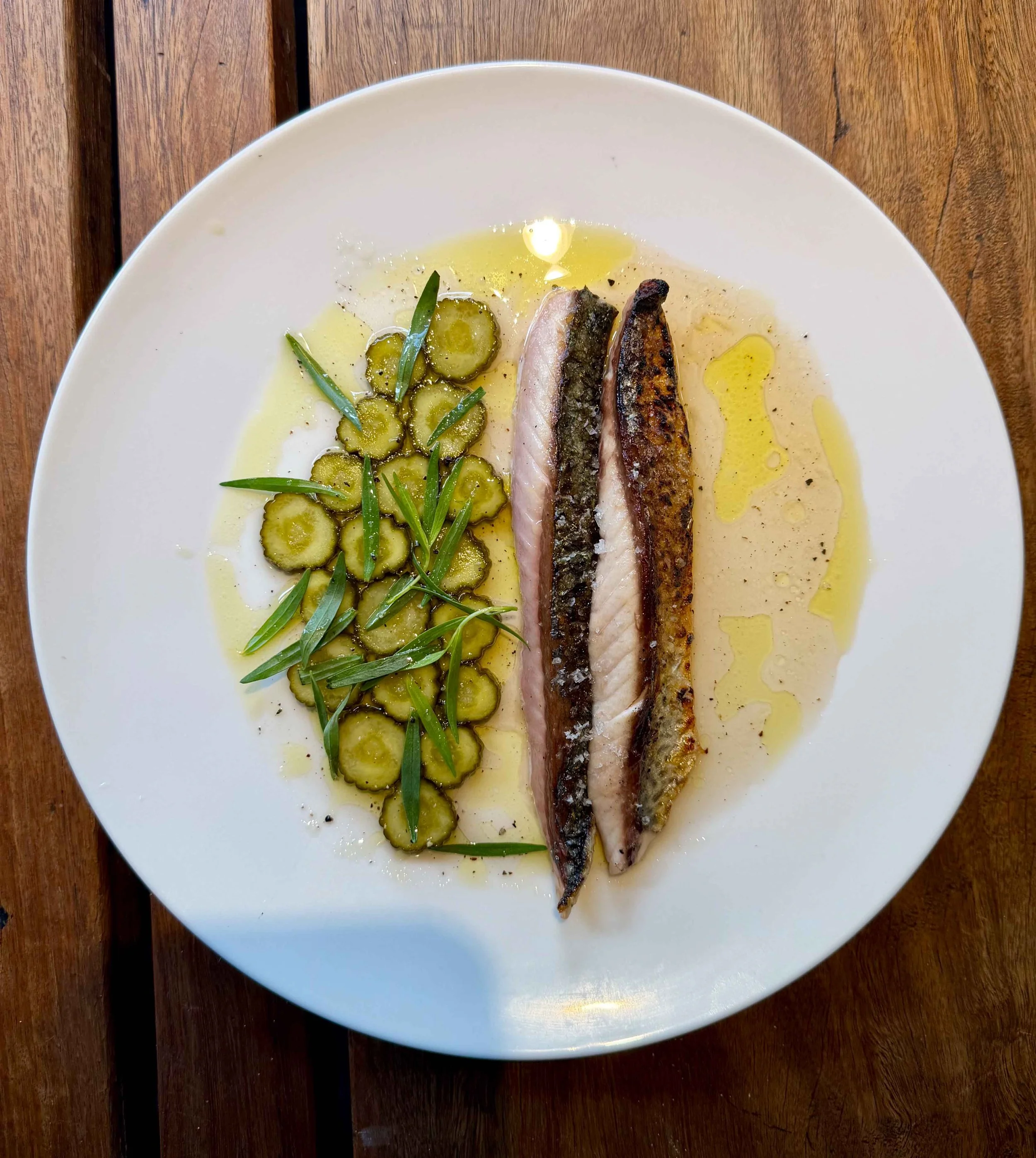 A white plate with grilled fish fillets and sliced green cucumbers garnished with fresh herbs, drizzled with olive oil, on a wooden surface.