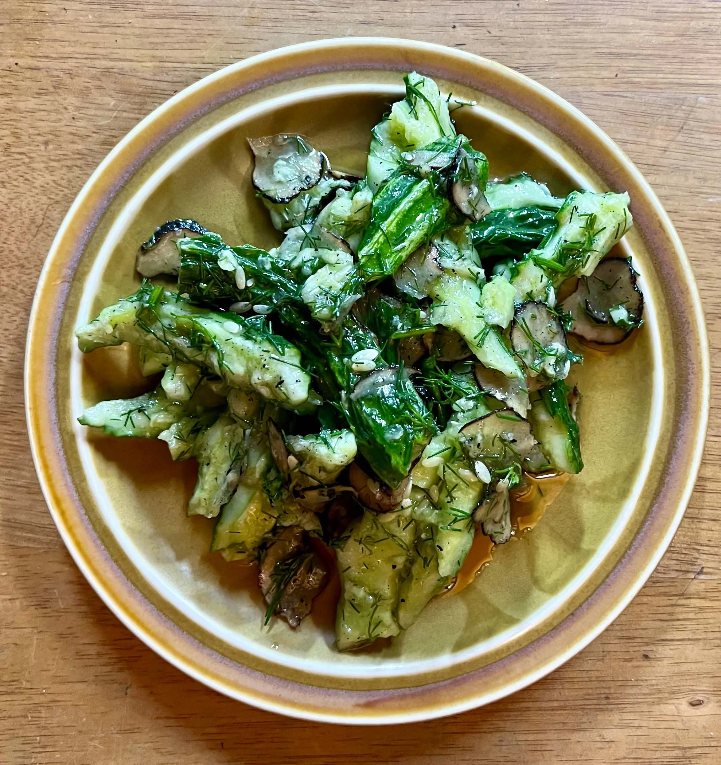 A ceramic bowl with a yellow and purple rim filled with a vegetable salad containing various greens, cucumbers, eggplant, and herbs, placed on a wooden surface.