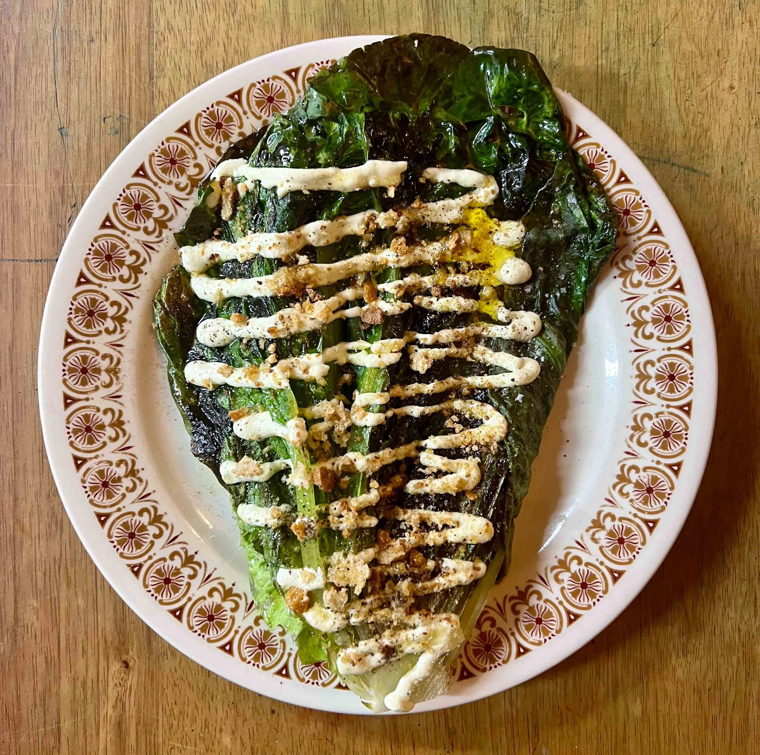 A dish of cooked greens topped with white sauce and sprinkled with crushed nuts, served on a decorative white plate on a wooden table.