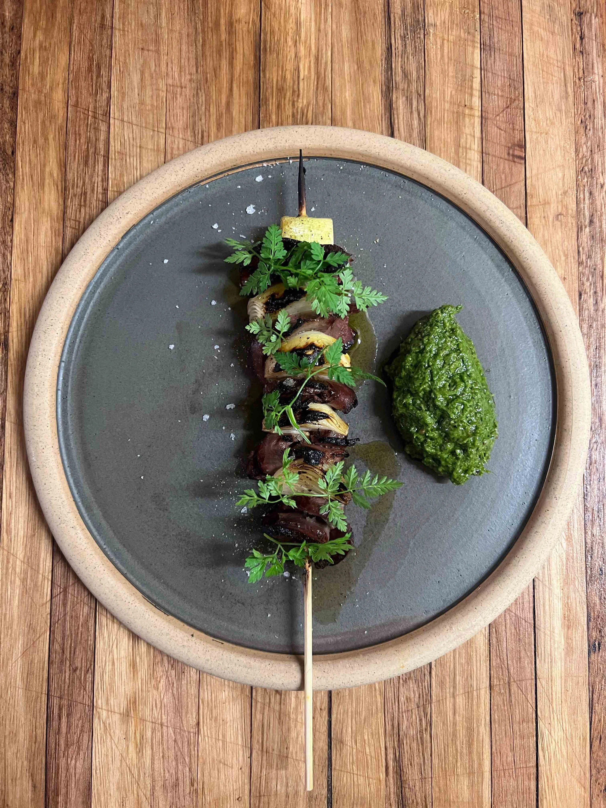 Skewered grilled meat and vegetables served on a dark plate with a green sauce, garnished with herbs, on a wooden surface.