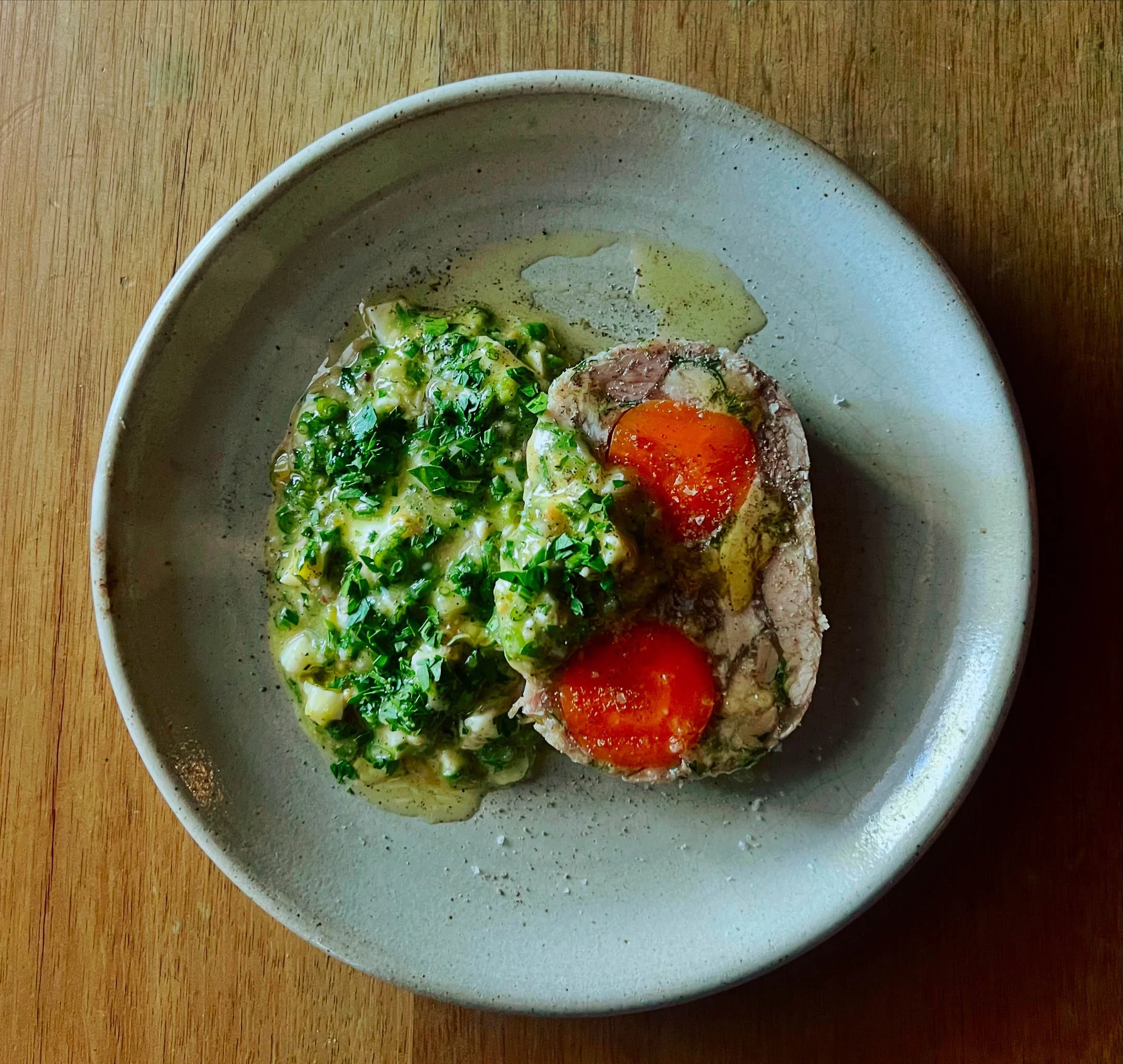 A ceramic bowl containing a dish with a baked egg, topped with a slice of tomato, served with a side of chopped green onions and herbs, all on a wooden table.