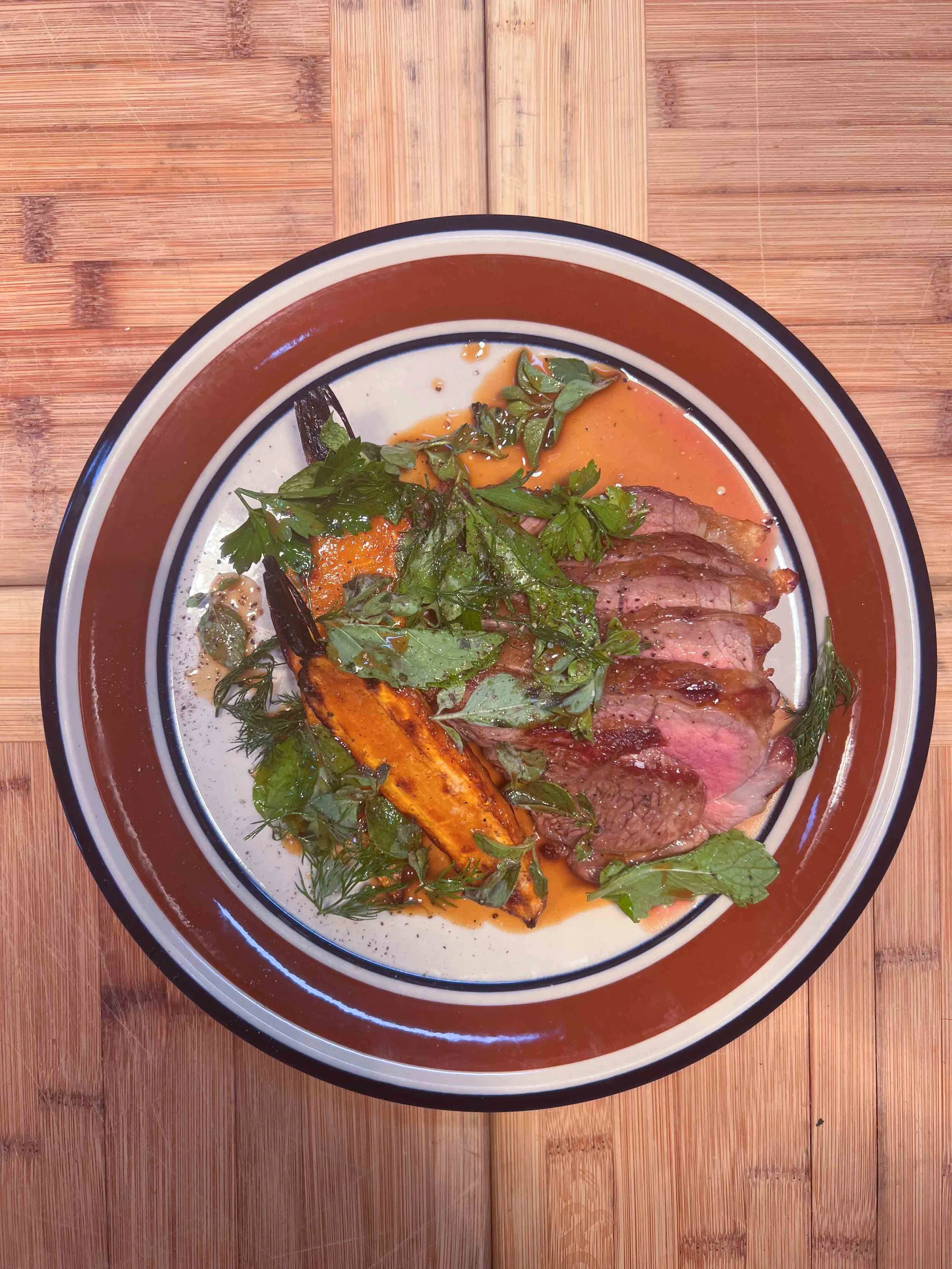 A bowl of steak slices with roasted vegetables and fresh green herbs on top, served in a brown and white ceramic dish on a wooden table.