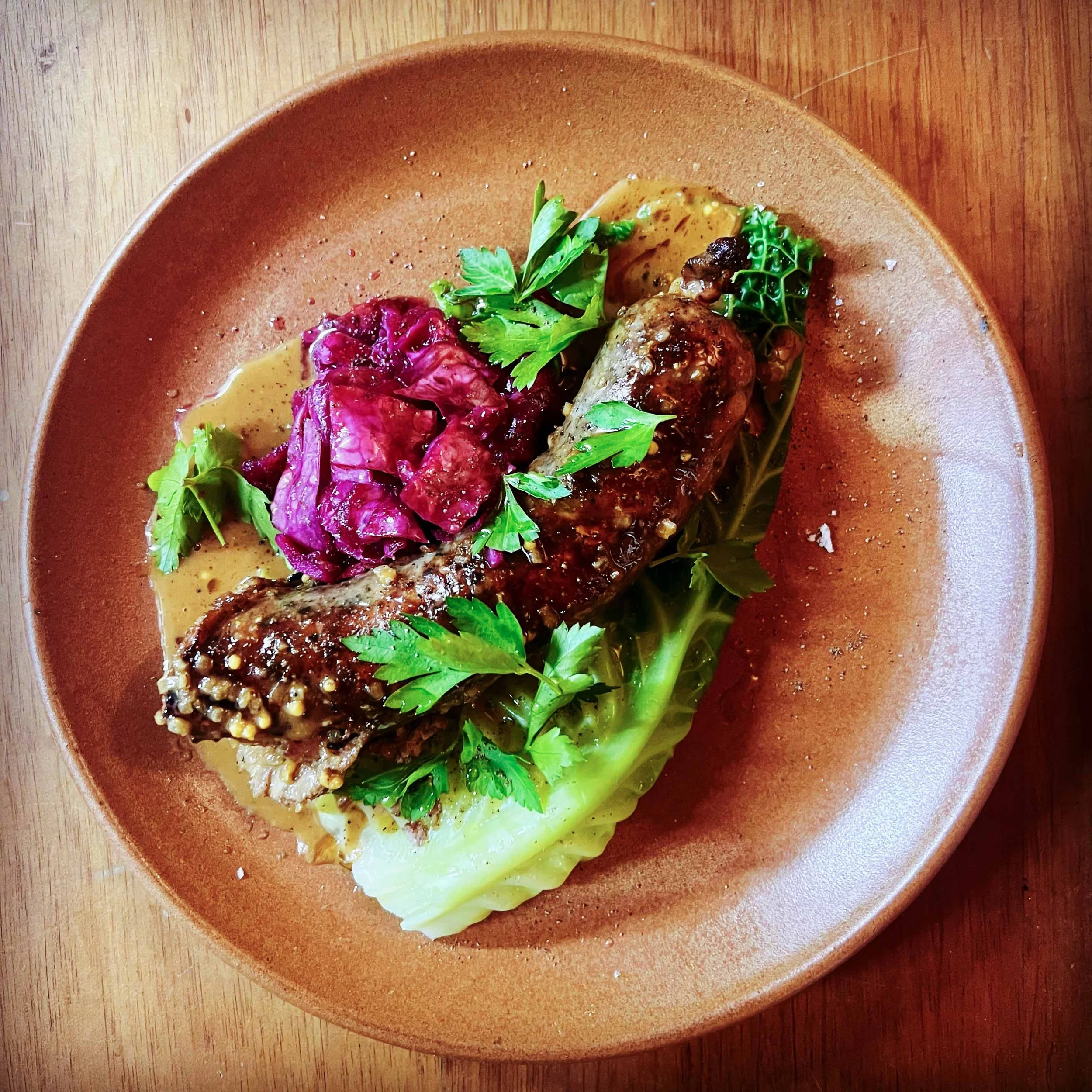 A plate of cooked chicken topped with sauce, garnished with fresh herbs, served alongside red cabbage and greens on a brown dish. The plate is on a wooden surface.