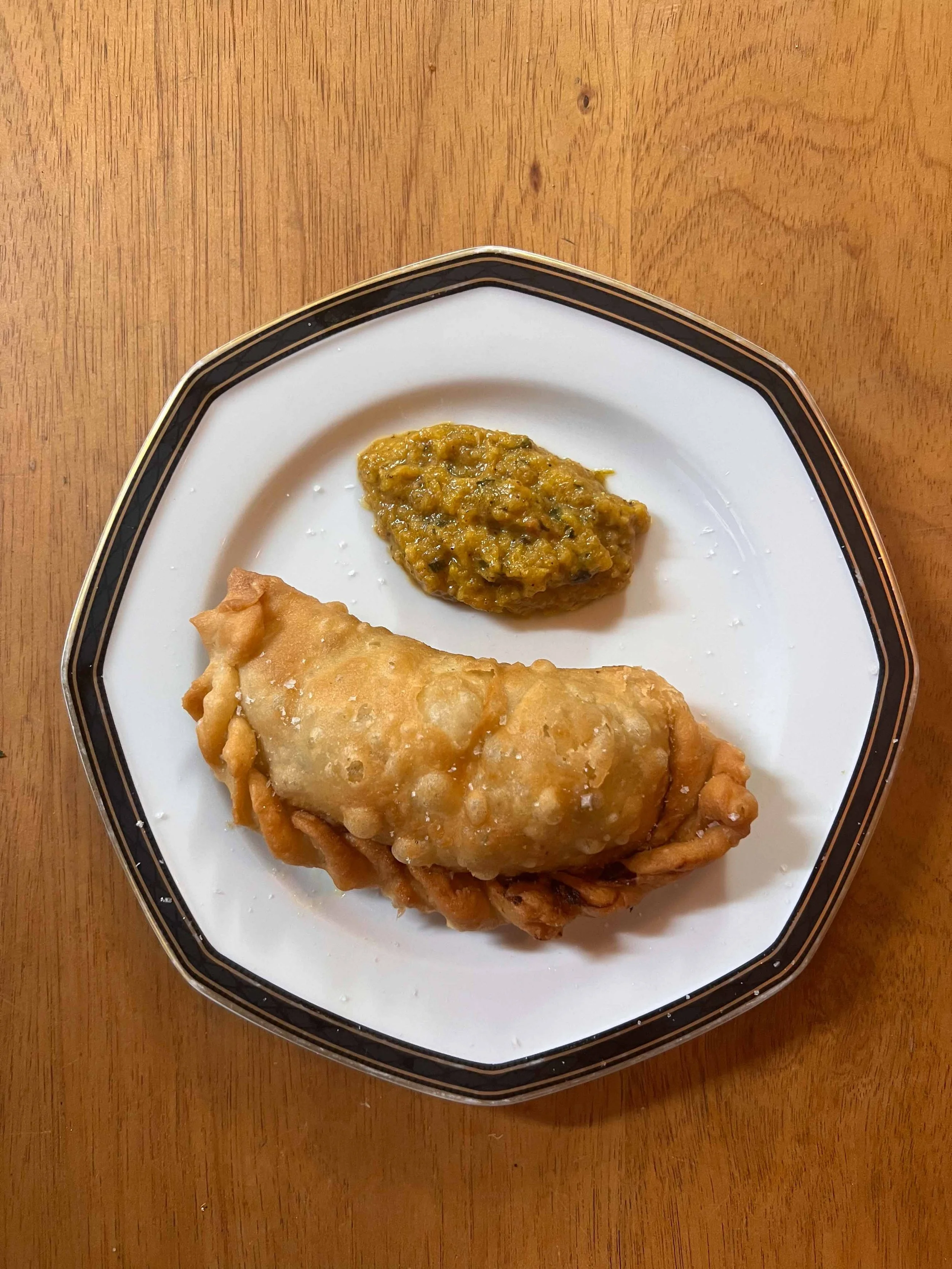 A white plate with a black and gold rim holding an Indian samosa and a serving of green chutney, placed on a wooden table.