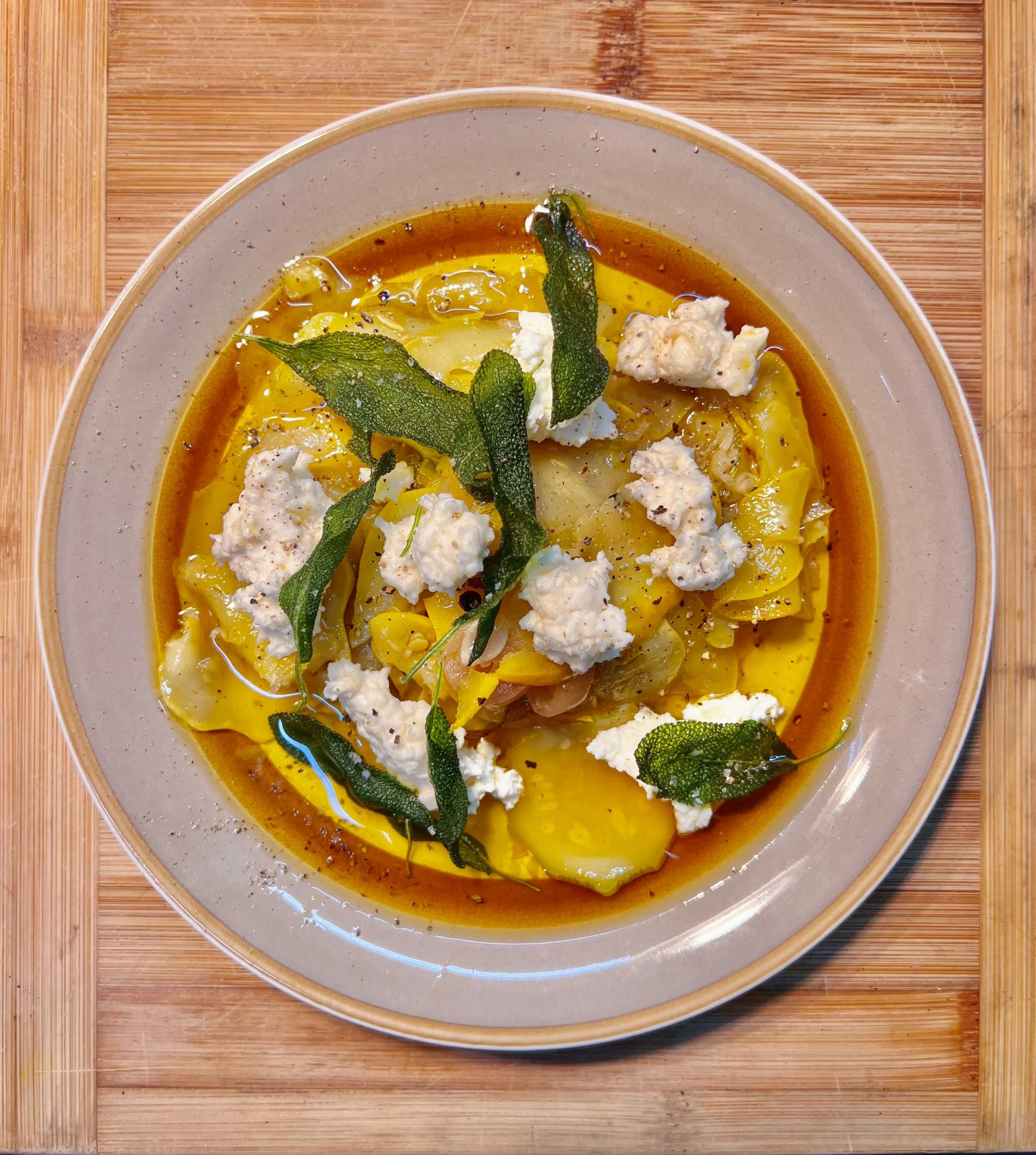 A bowl of pumpkin or squash soup garnished with fresh sage leaves, ricotta cheese, and black pepper, placed on a wooden surface.