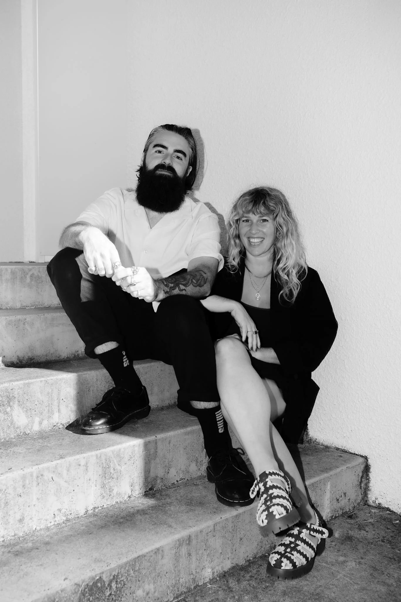 A black and white photo of a man with a beard and long hair and a woman with curly hair sitting on concrete stairs.