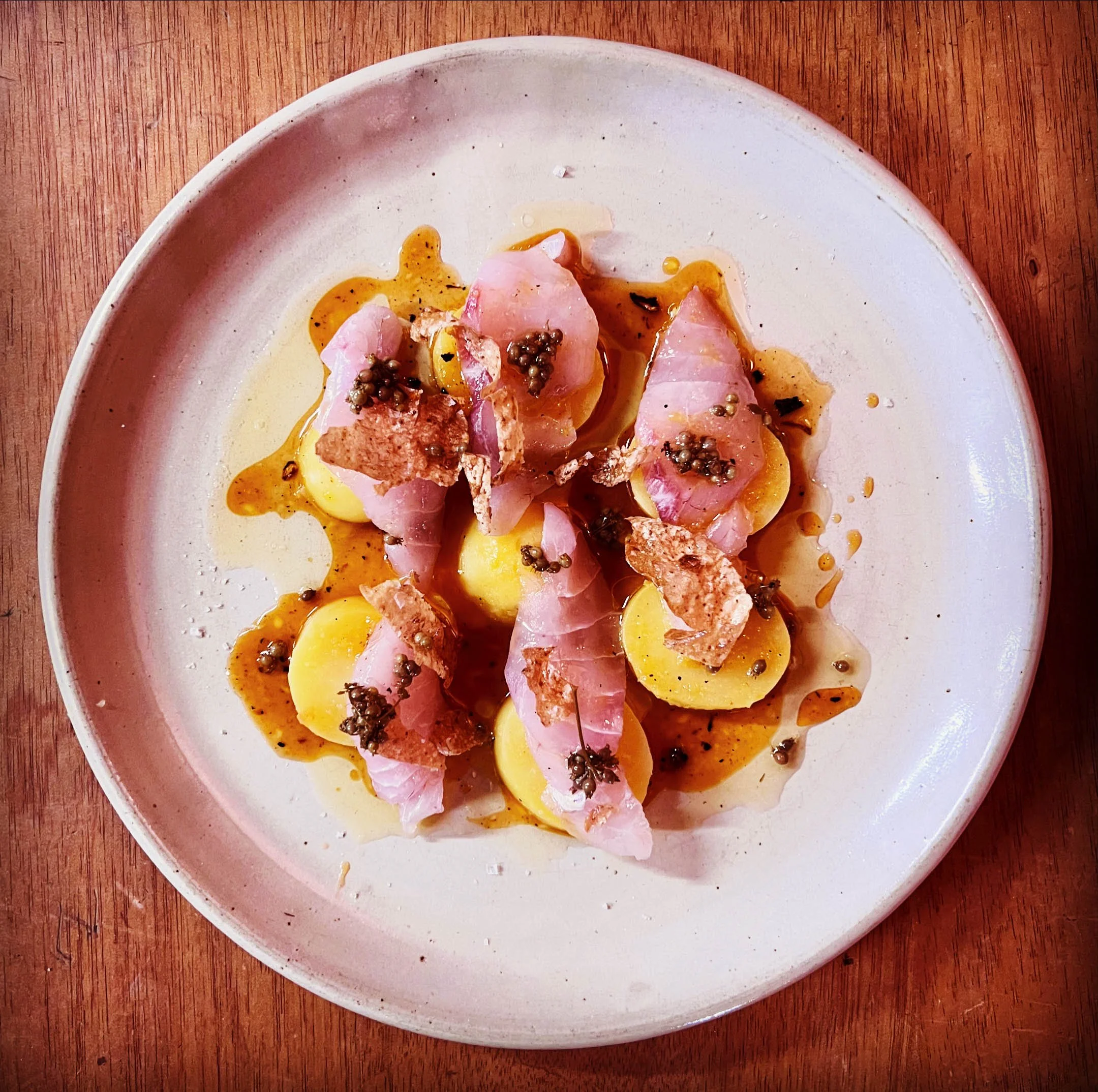 Sliced ham and yellow potatoes garnished with black pepper and mustard seeds on a white oval plate, placed on a wooden table.