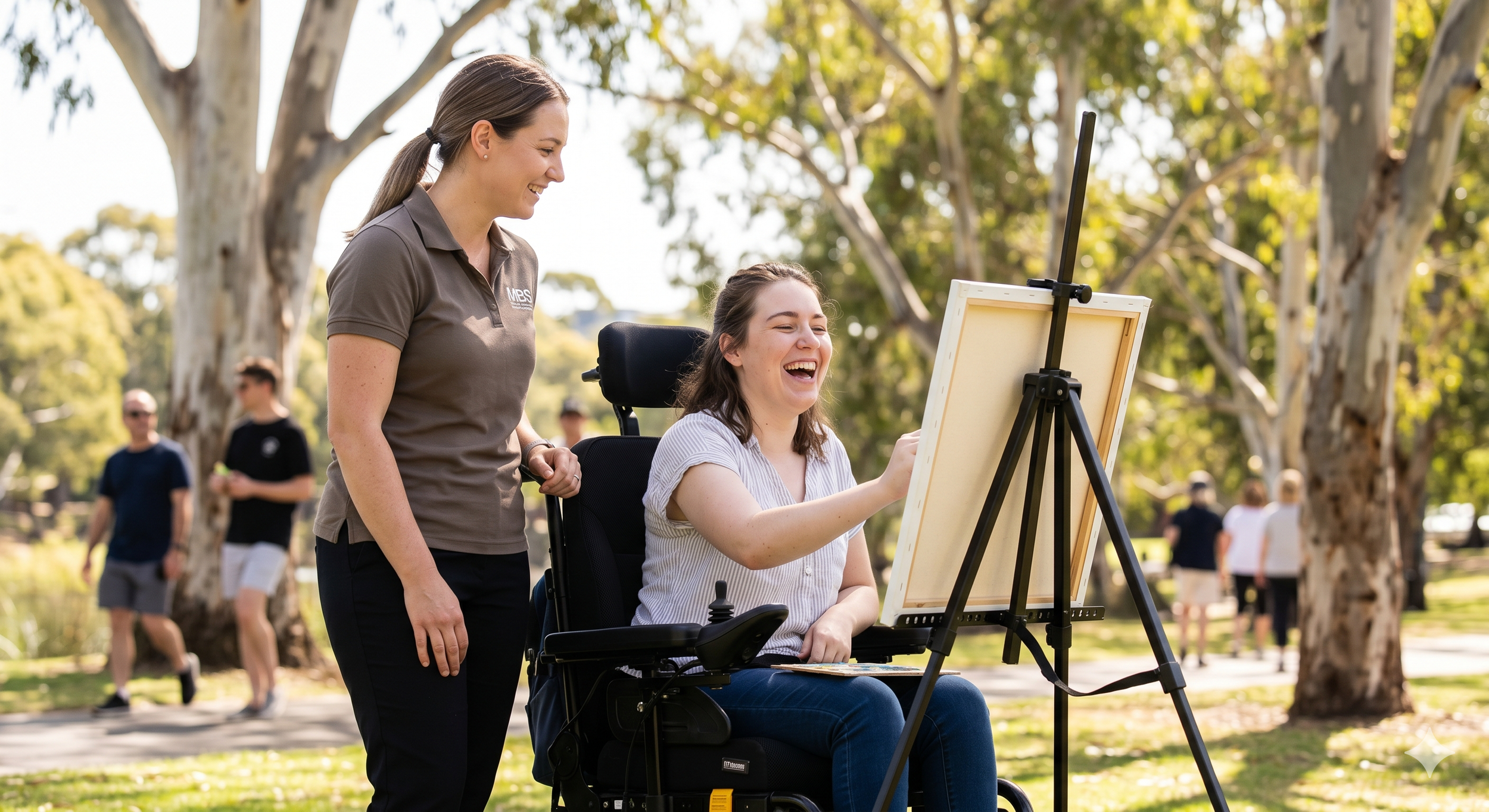 A woman in a brown shirt smiling and standing next to a young woman in a wheelchair who is drawing or painting on a canvas set up outdoors among trees, with several people walking in the background.