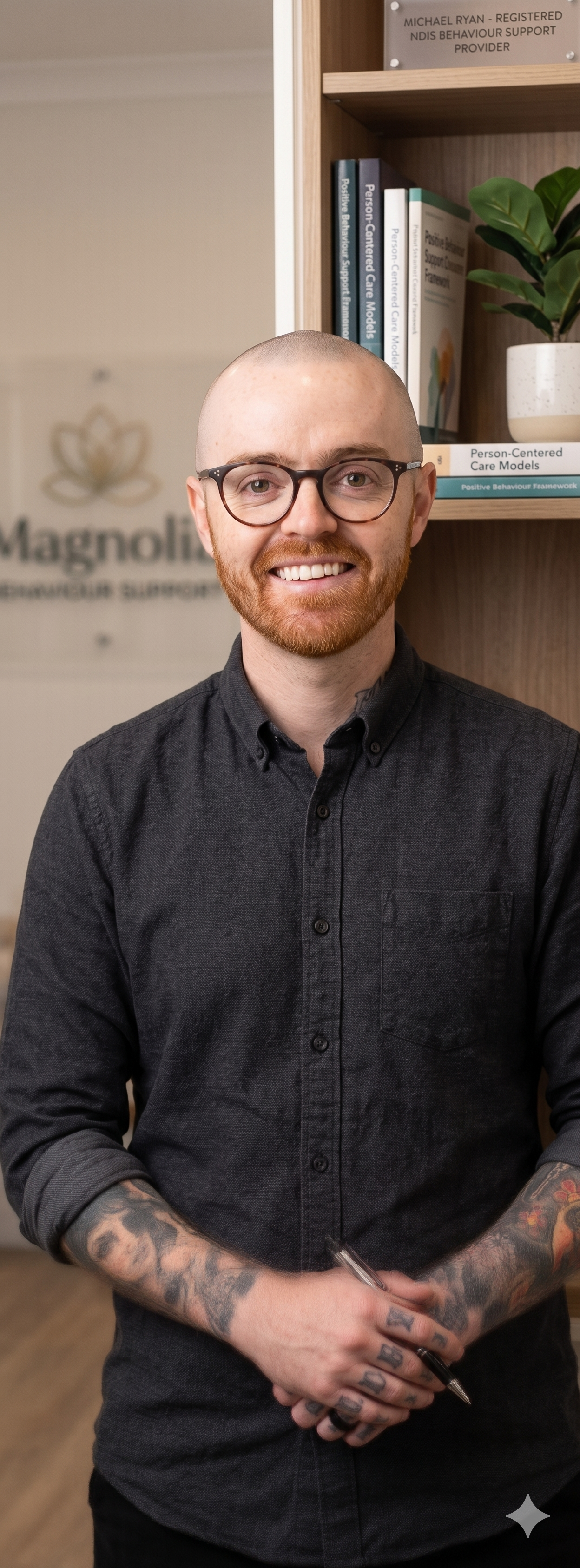 Portrait of a young man with glasses, a red beard, and tattoos, smiling and holding a pen, standing in an office or clinic with shelves of books and a potted plant behind him.