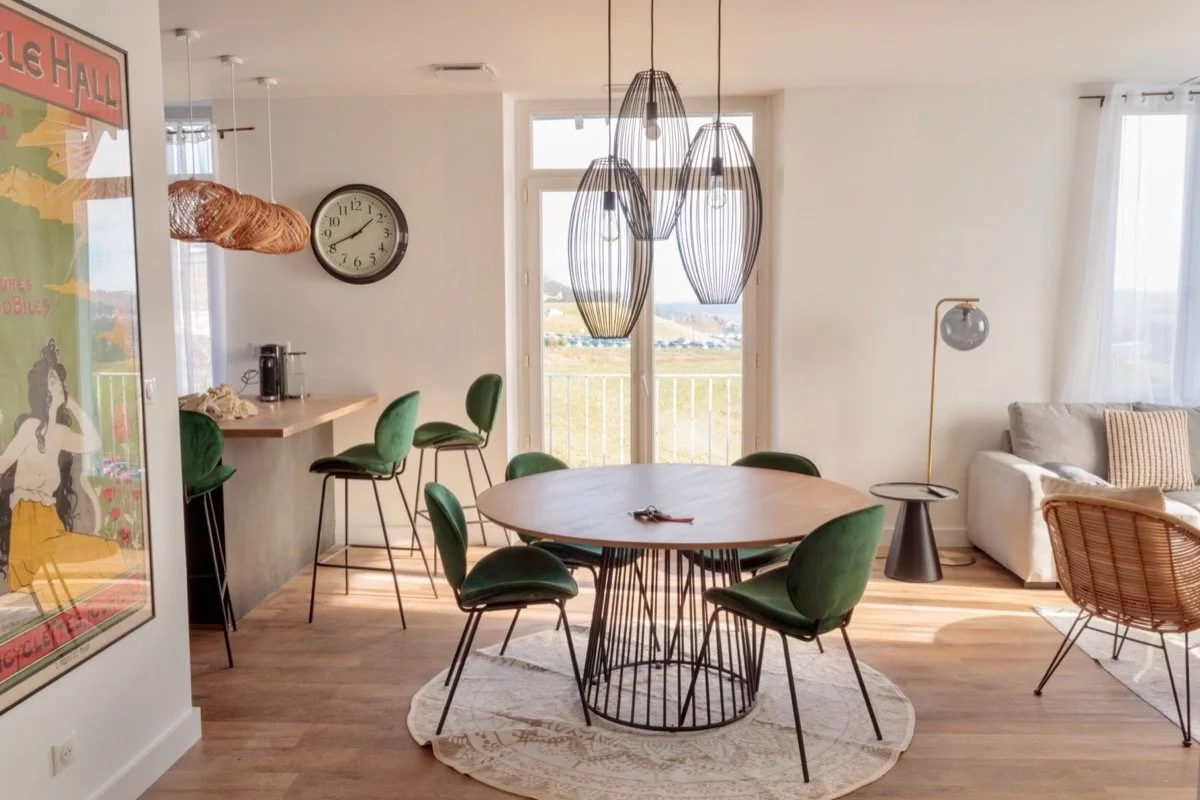 Salon moderne avec une table ronde en bois, six chaises en velours vert, un meuble en rotin, un canapé beige, une lampe de lecture et une fresque d'art dans une pièce lumineuse et décorée de manière contemporaine.