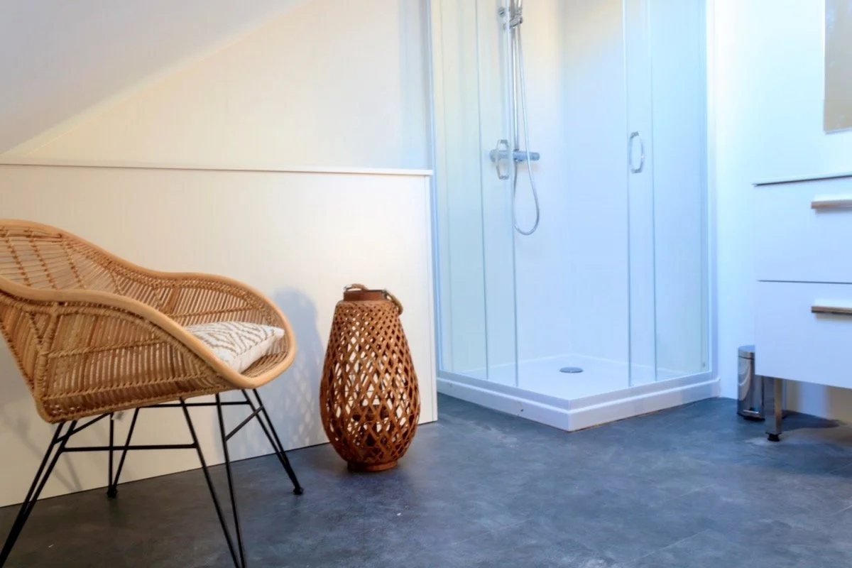 Une salle de bain avec une cabine de douche en verre, un meuble blanc avec tiroirs, une chaise en rotin, une lampe décorative en rotin et un sol en carrelage gris.