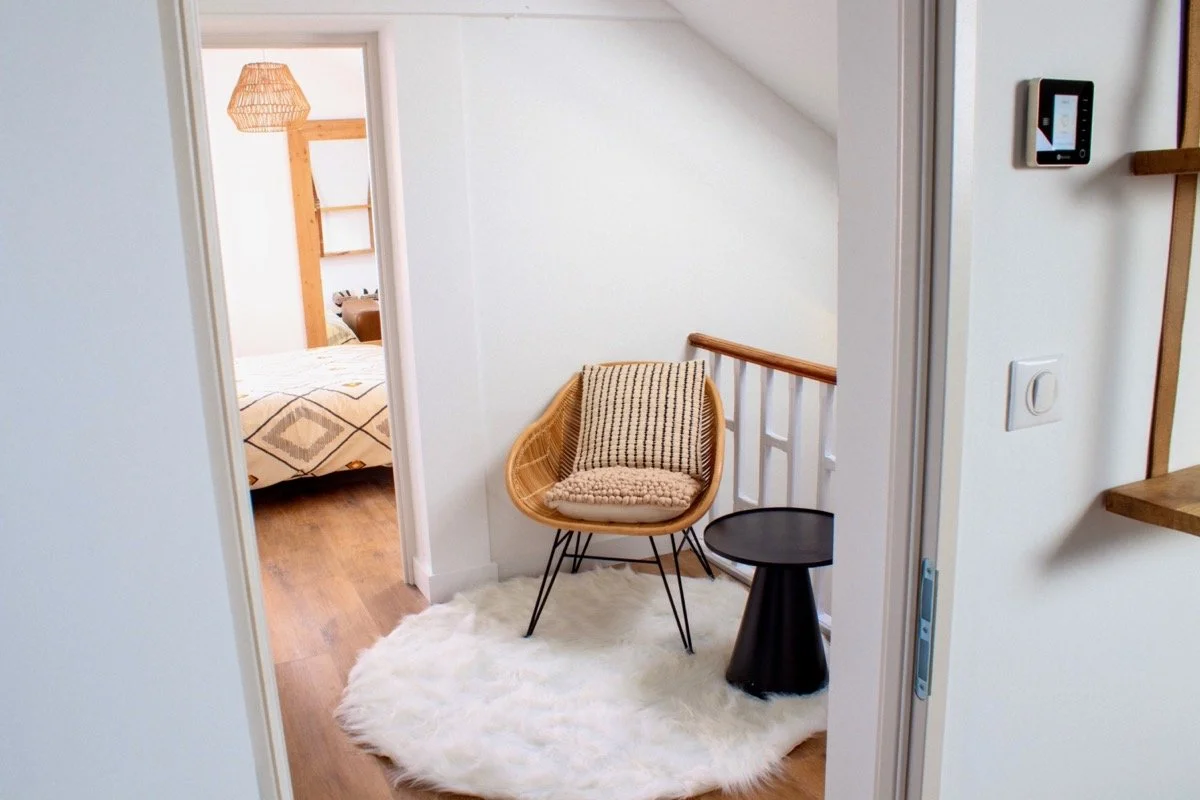 Espace de coin lecture avec fauteuil en rotin, coussin et petit table noire, tapis blanc, dans un appartement moderne.