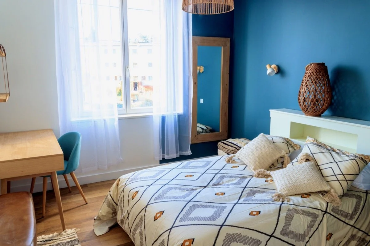 Chambre avec un lit, des coussins, une grande fenêtre avec des rideaux blancs, un miroir en bois au mur, une table en bois, et une lampe en rotin