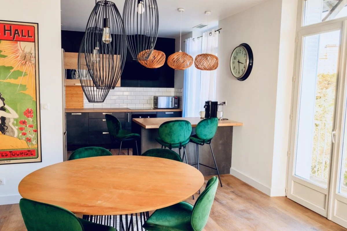 Une cuisine et salle à manger modernes avec un tableau vintage sur le mur, une table ronde en bois avec des chaises vertes, un bar avec des chaises vertes, des lampes suspendues en osier et un mur blanc avec une horloge.