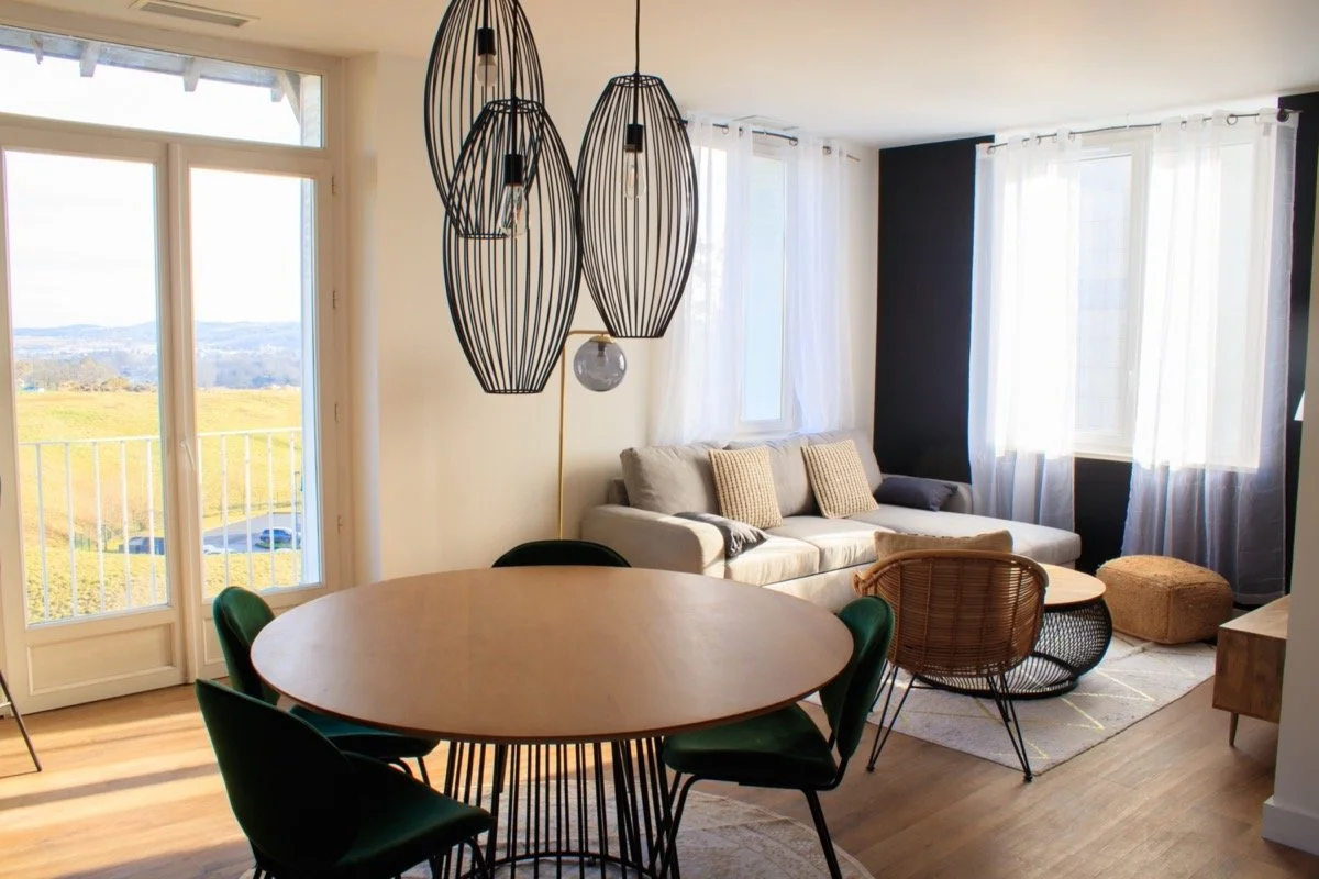 Salon et salle à manger modernes avec grand canapé beige, fauteuils en rotin, table ronde en bois, lampes suspendues en métal noir, grandes fenêtres avec rideaux blancs, vue sur un paysage extérieur.
