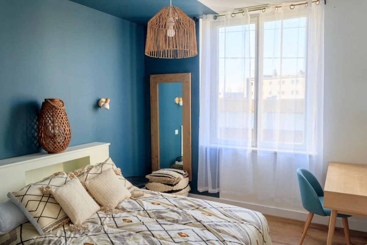 Chambre moderne avec mur bleu, lit avec literie beige et coussins, miroir, lampes, grande fenêtre avec rideaux blancs, lampe en rotin