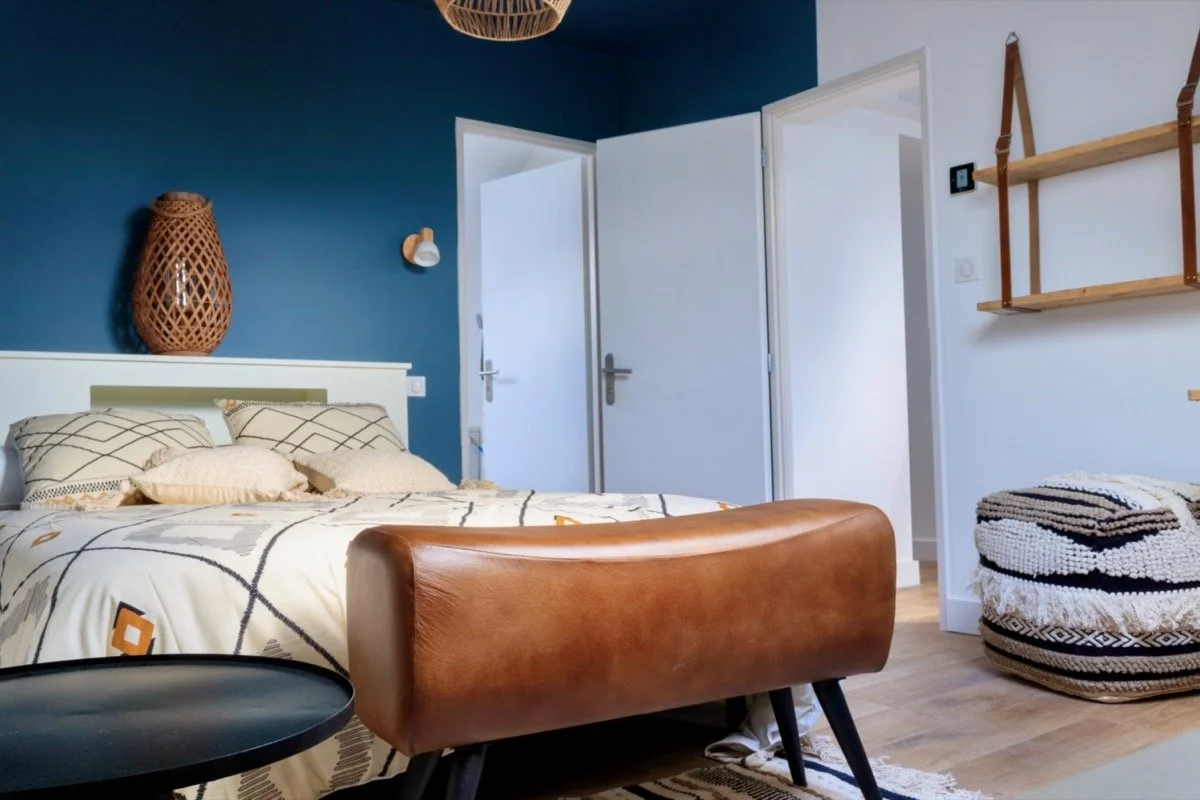 Chambre moderne avec lit à tête blanche, murs bleu, porte blanche, étagère en bois suspendue, pouf tissé et plante en jarre.