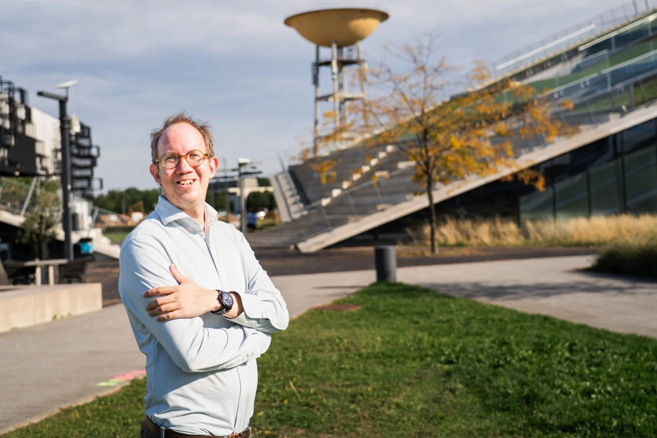 Michaël staat buiten met moderne architectuur en een boom met gele bladeren op de achtergrond, glimlachend en met armen gekruist.