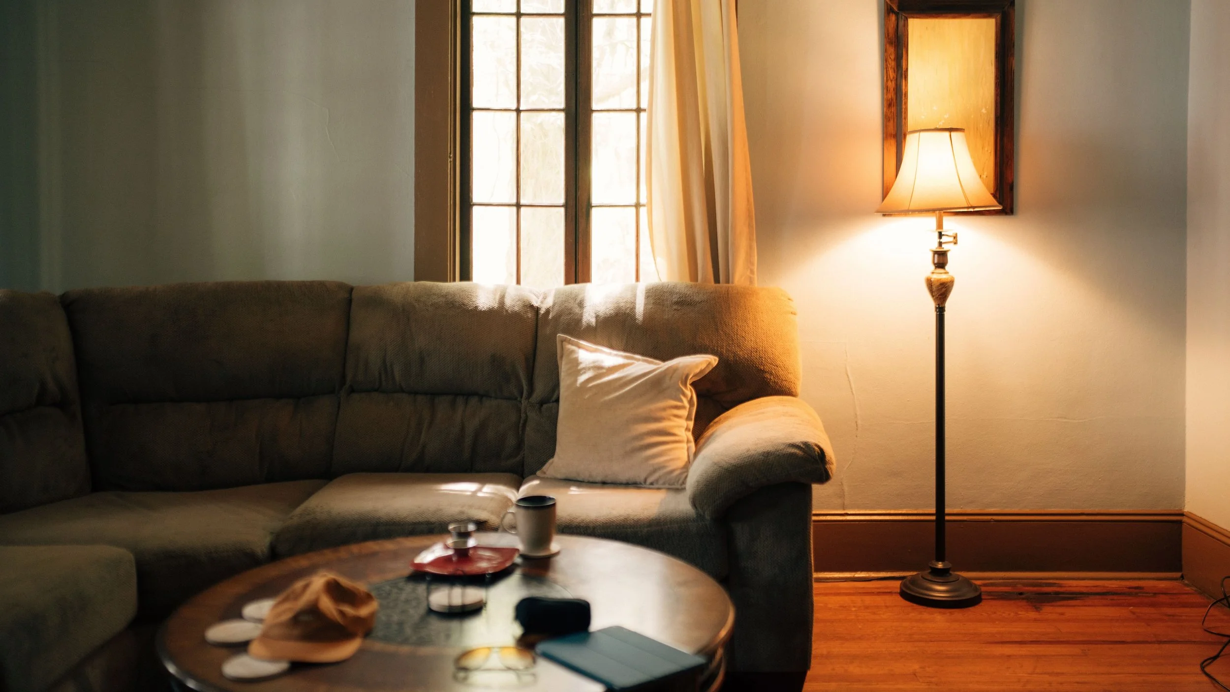 A cozy living room corner with a brown sofa, a white pillow on it, and a wooden coffee table with various items including a mug and a hat. In the background, there is a window with curtains and a tall floor lamp with a beige shade, illuminating the space.