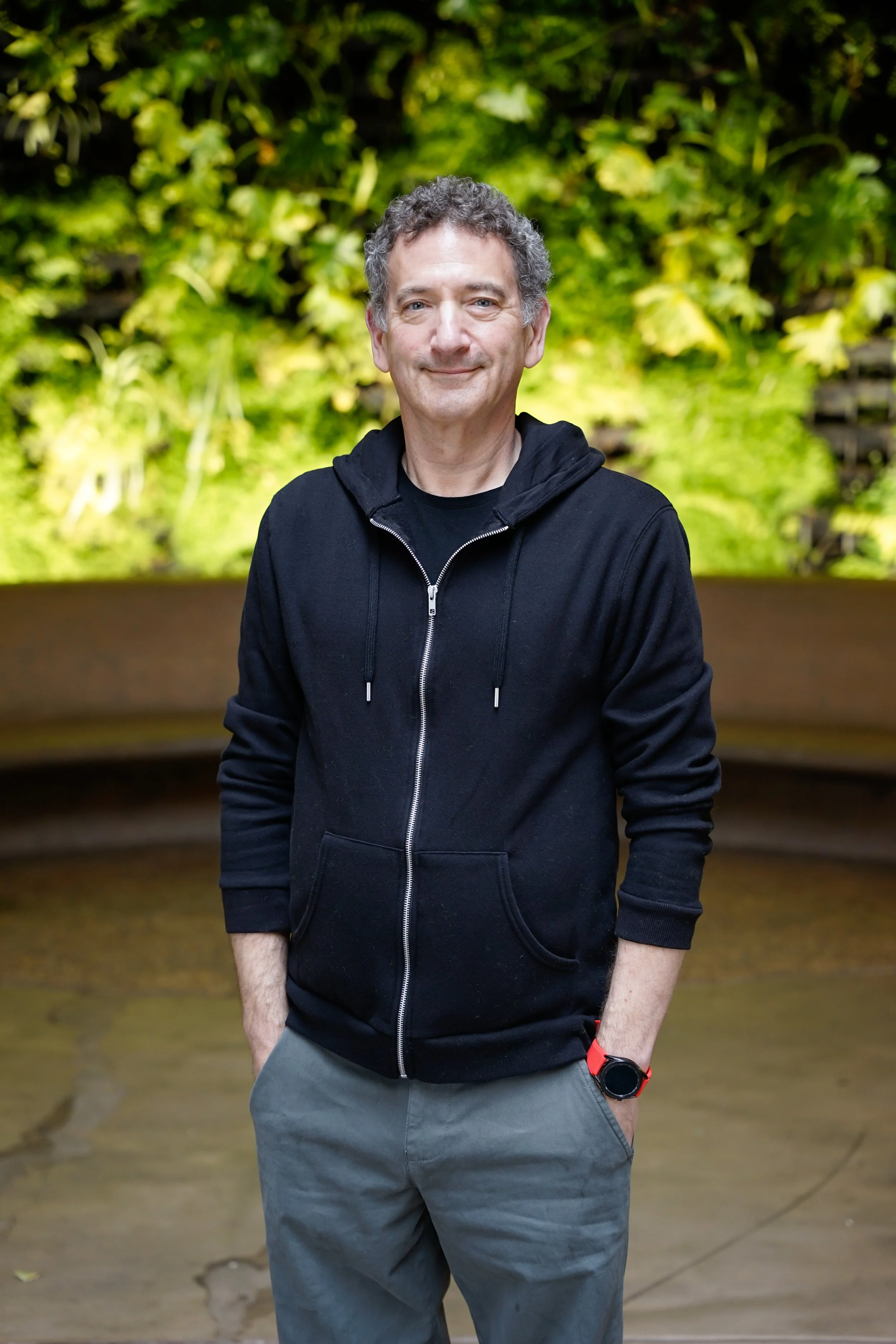 A middle-aged man with curly gray hair, wearing a black zip-up hoodie, gray pants, and a red smartwatch, standing outdoors in front of a background of lush green foliage, smiling with his hands in his pockets.