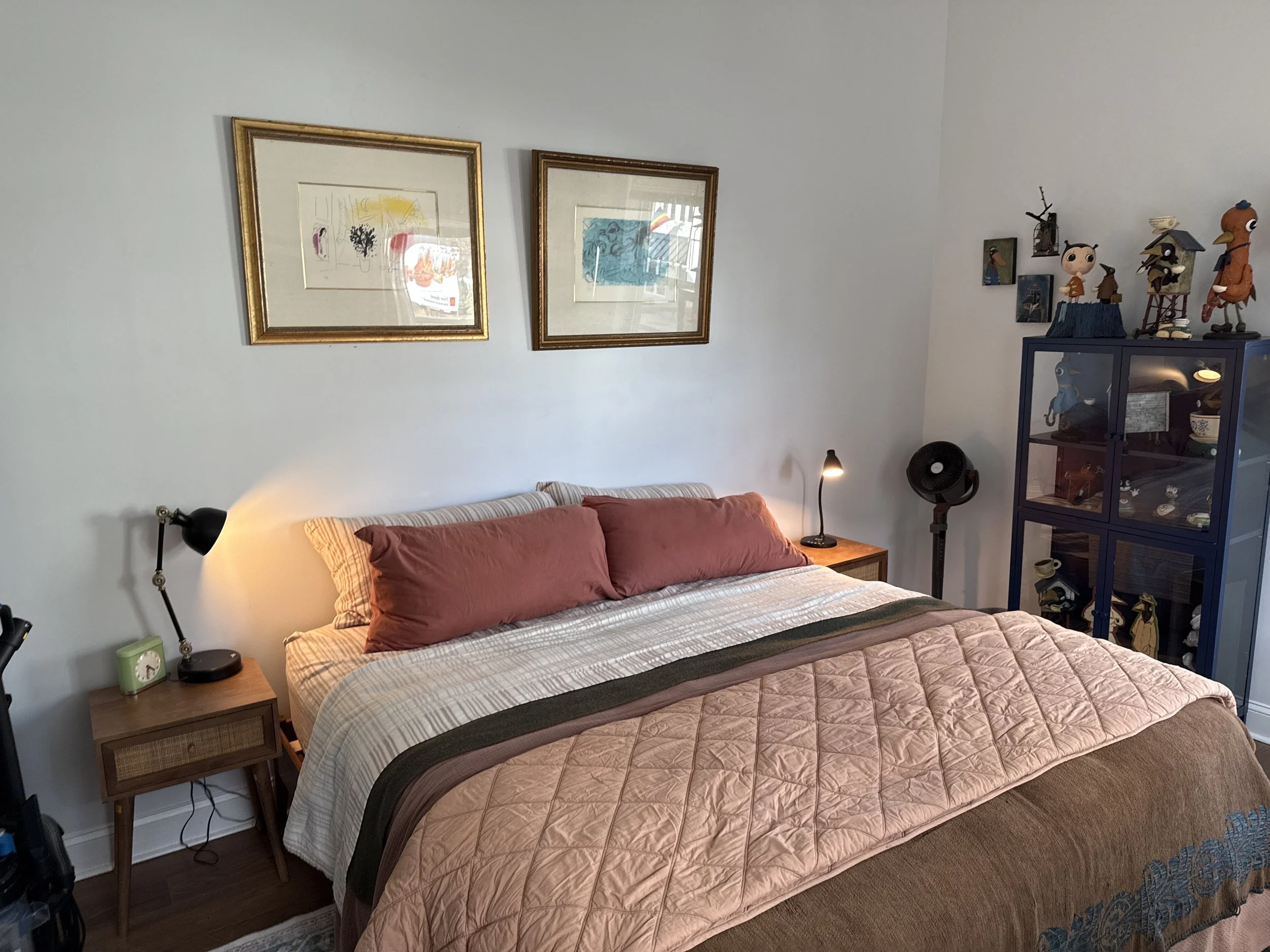 A cozy bedroom with a bed covered in a beige quilt and two pinkish-red pillows. To the left, there is a nightstand with a green clock and a black desk lamp. Behind the bed, a white wall features two framed abstract art pieces. On the right side, a dark blue display cabinet showcases various collectibles, with small artworks and figurines displayed on top. A black fan and a desk lamp are also visible in the room.