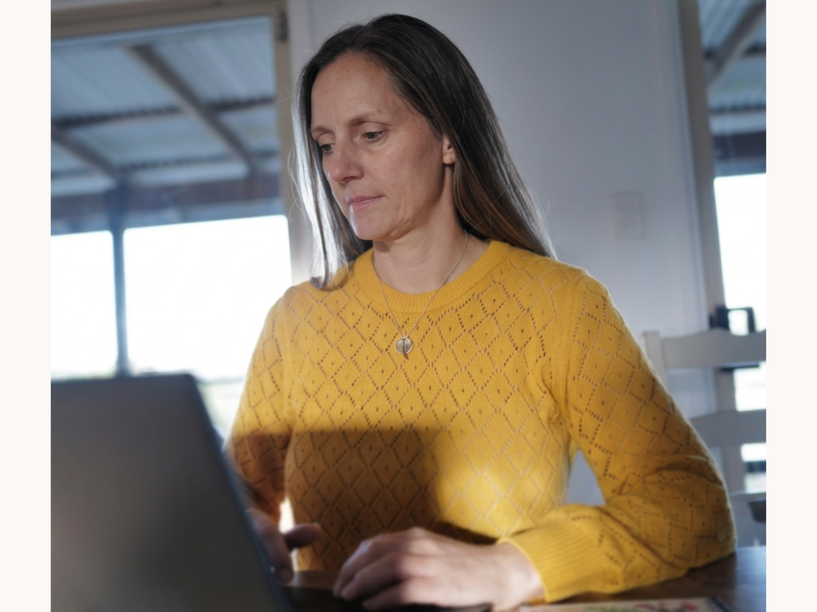 Christine in a yellow sweater, working on a laptop in a bright room with large windows.