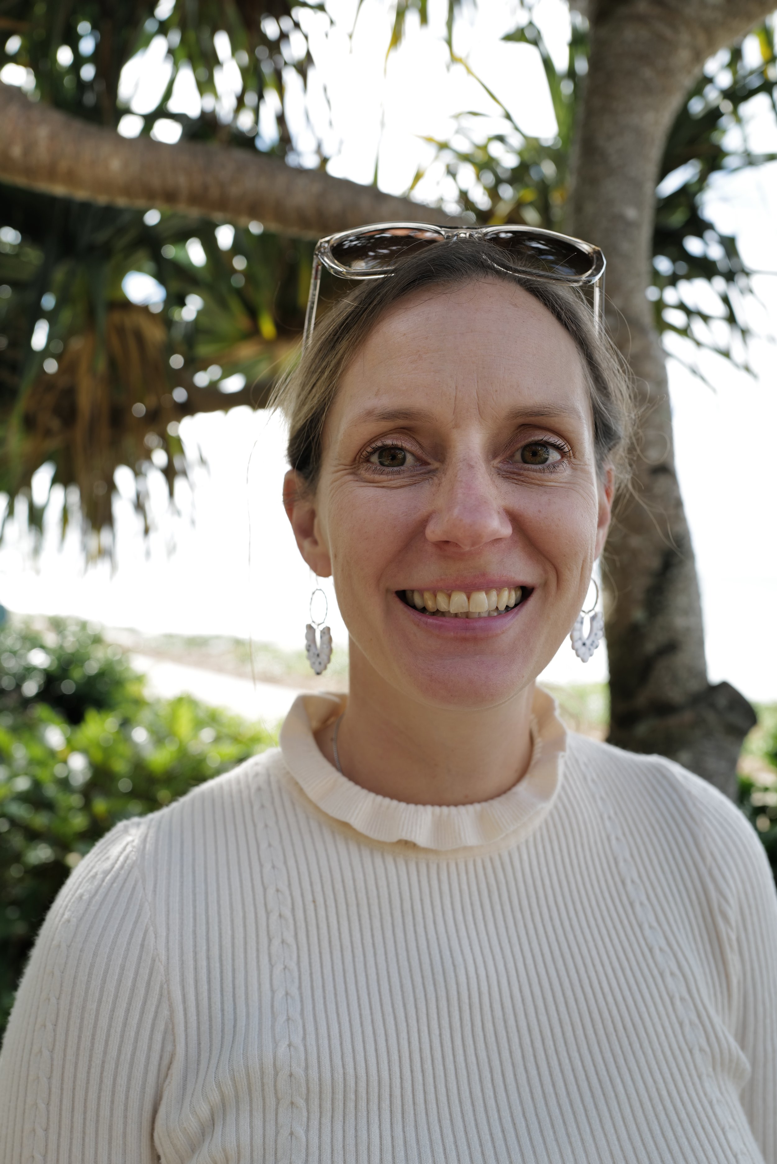 Christine - founder of Hinterland Marketing Co. smiling outdoors with a tree in the background, wearing sunglasses on her head, earrings, and a white ribbed sweater.