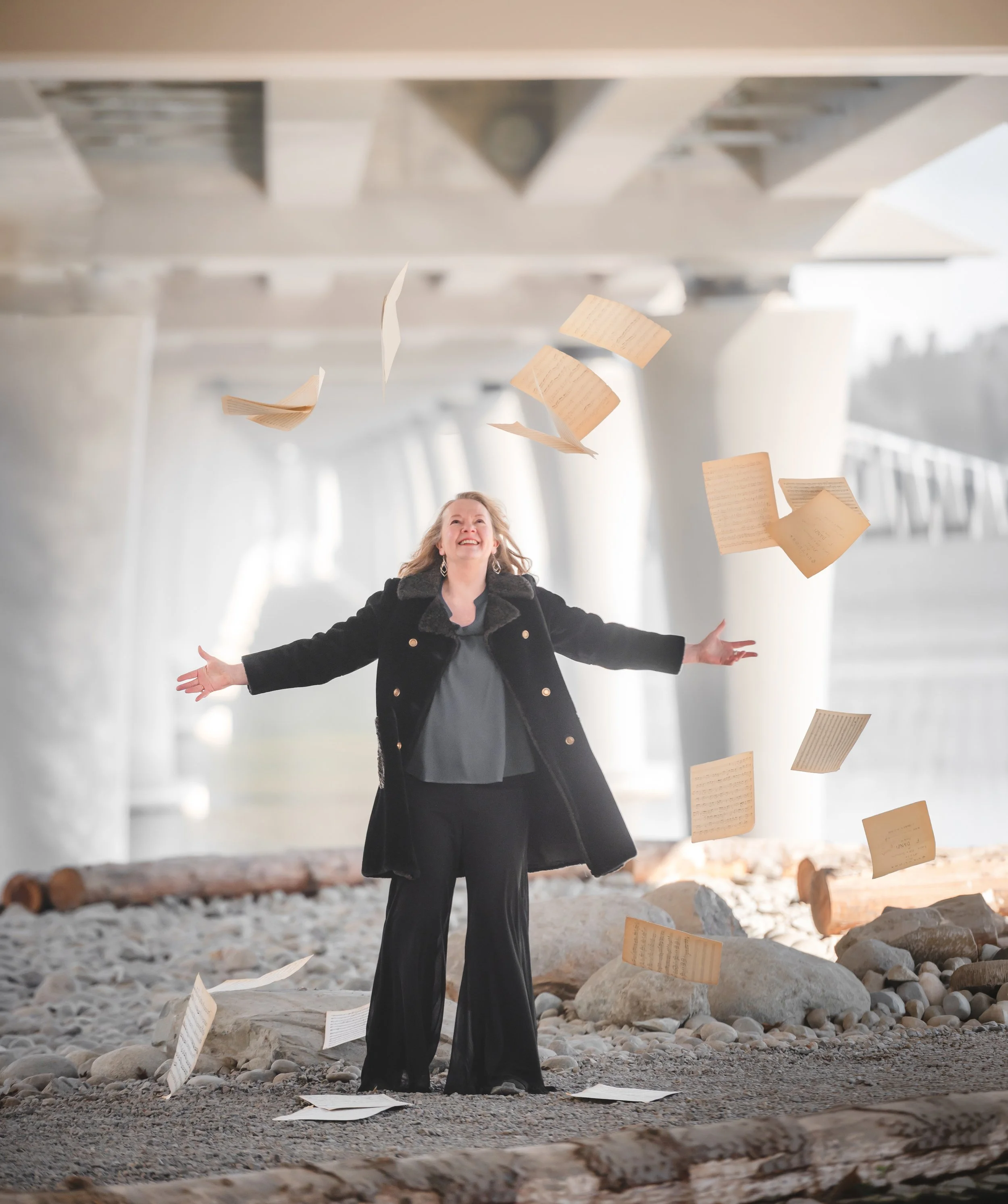 A woman standing outdoors under a bridge, with arms stretched out, smiling, with papers flying around her, some on the ground and objects like rocks and logs surrounding her.