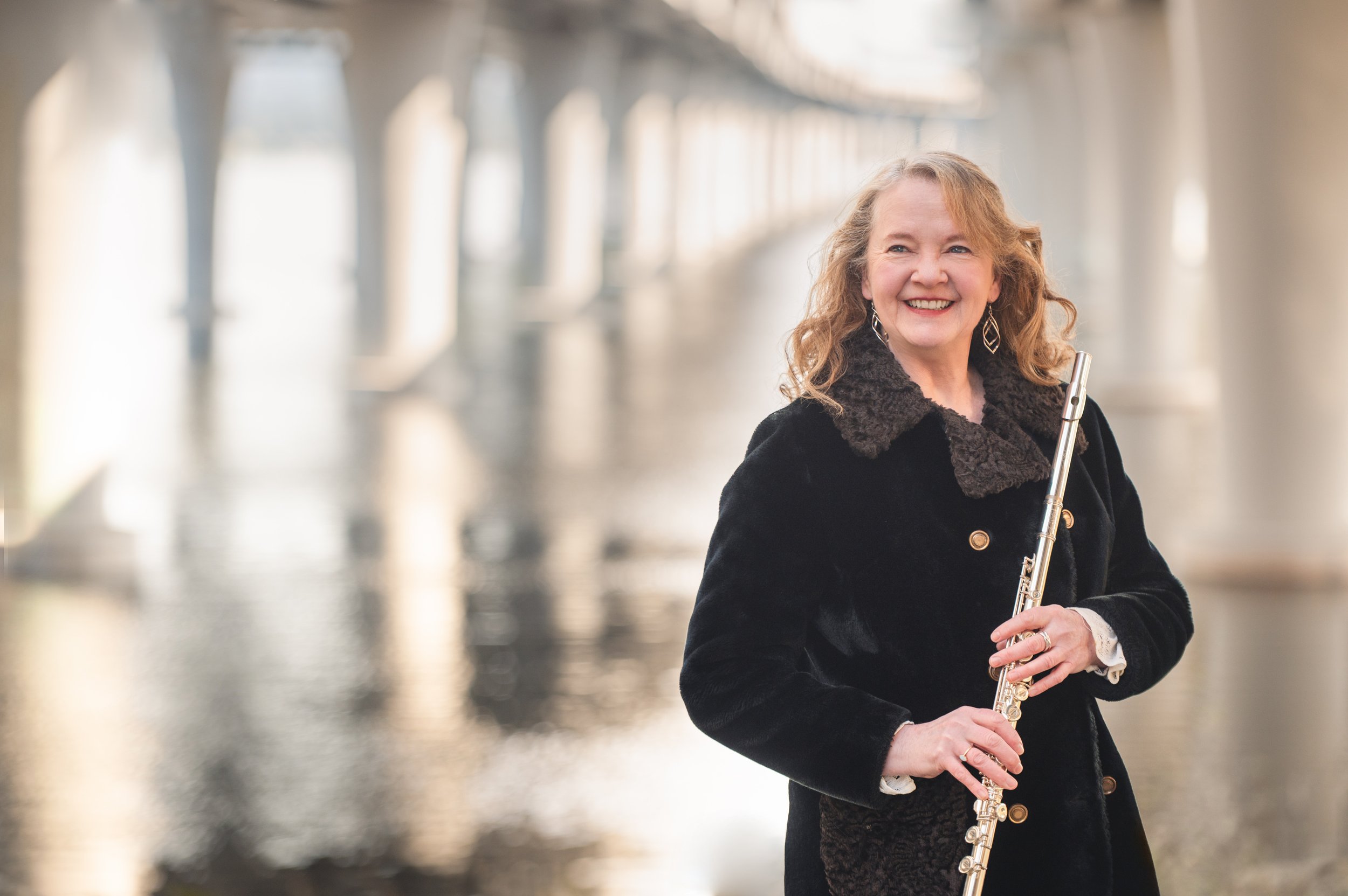 A woman with curly red hair playing a flute outdoors in front of a blurred background of a bridge and water.