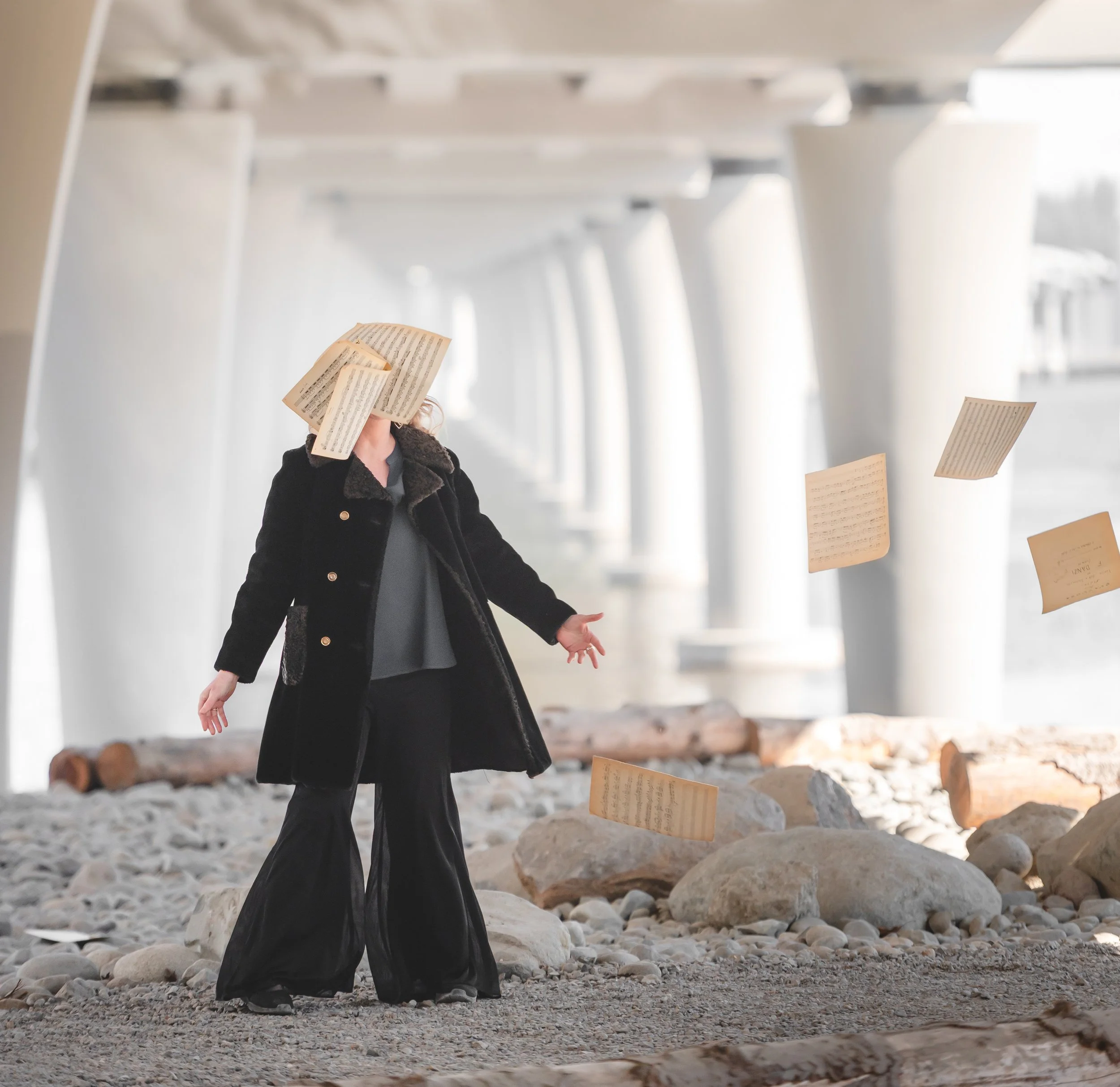 A person standing on rocky ground inside a large, open industrial or warehouse space with high ceiling and metal beams, holding a sheet of music or paper near their face, with other sheets of paper floating or suspended around them.
