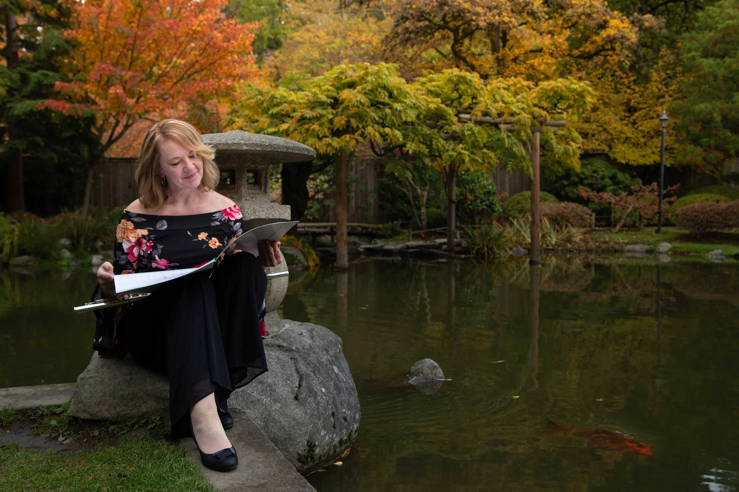 A woman with blonde hair wearing a floral off-the-shoulder top and black skirt, sitting on a rock by a pond in a garden with fall foliage, reading a book, with a flute resting on her lap.