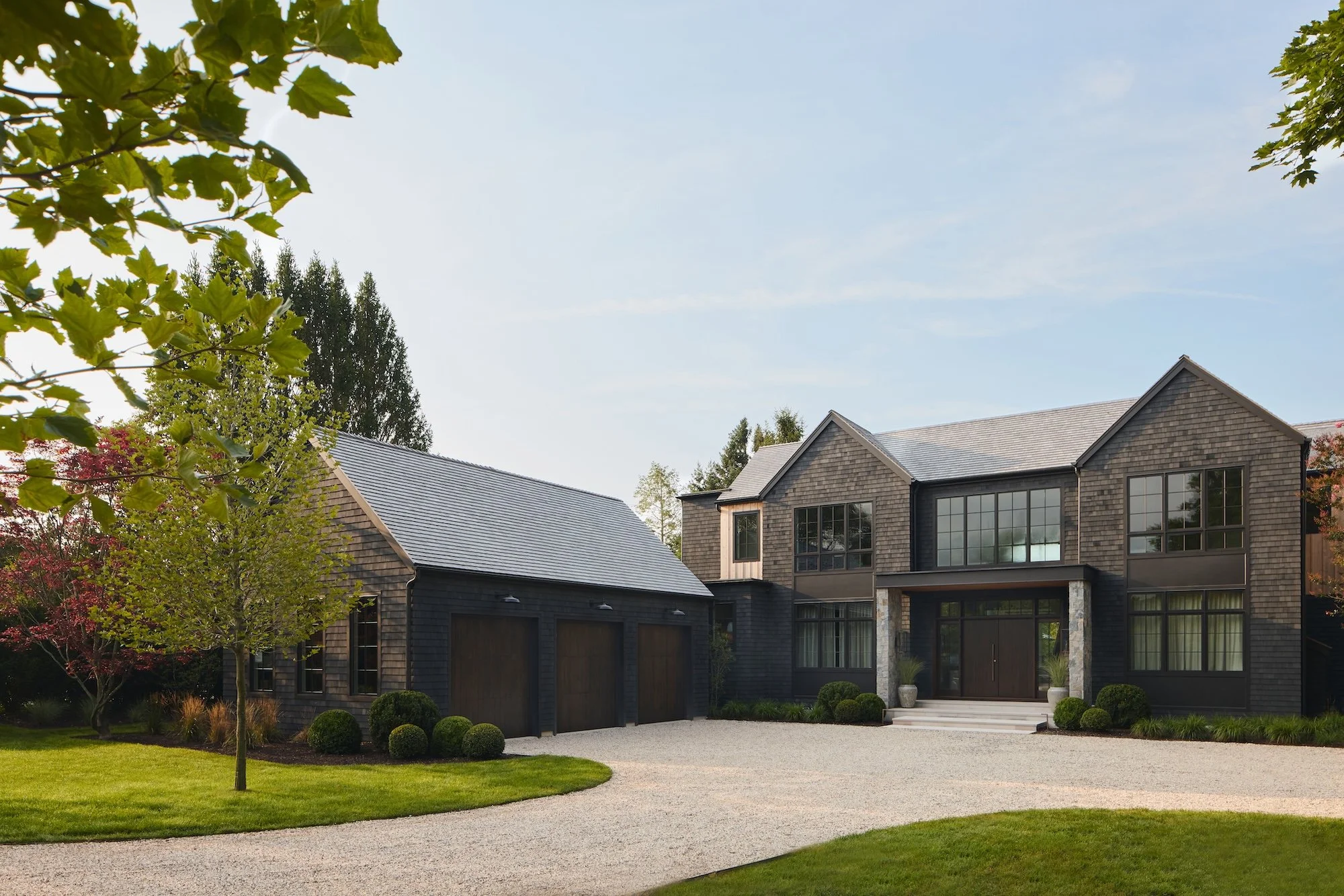 A modern residential house with a gray shingle exterior, large windows, and a garage, surrounded by a well-maintained lawn, trees, and a gravel driveway under a clear sky.
