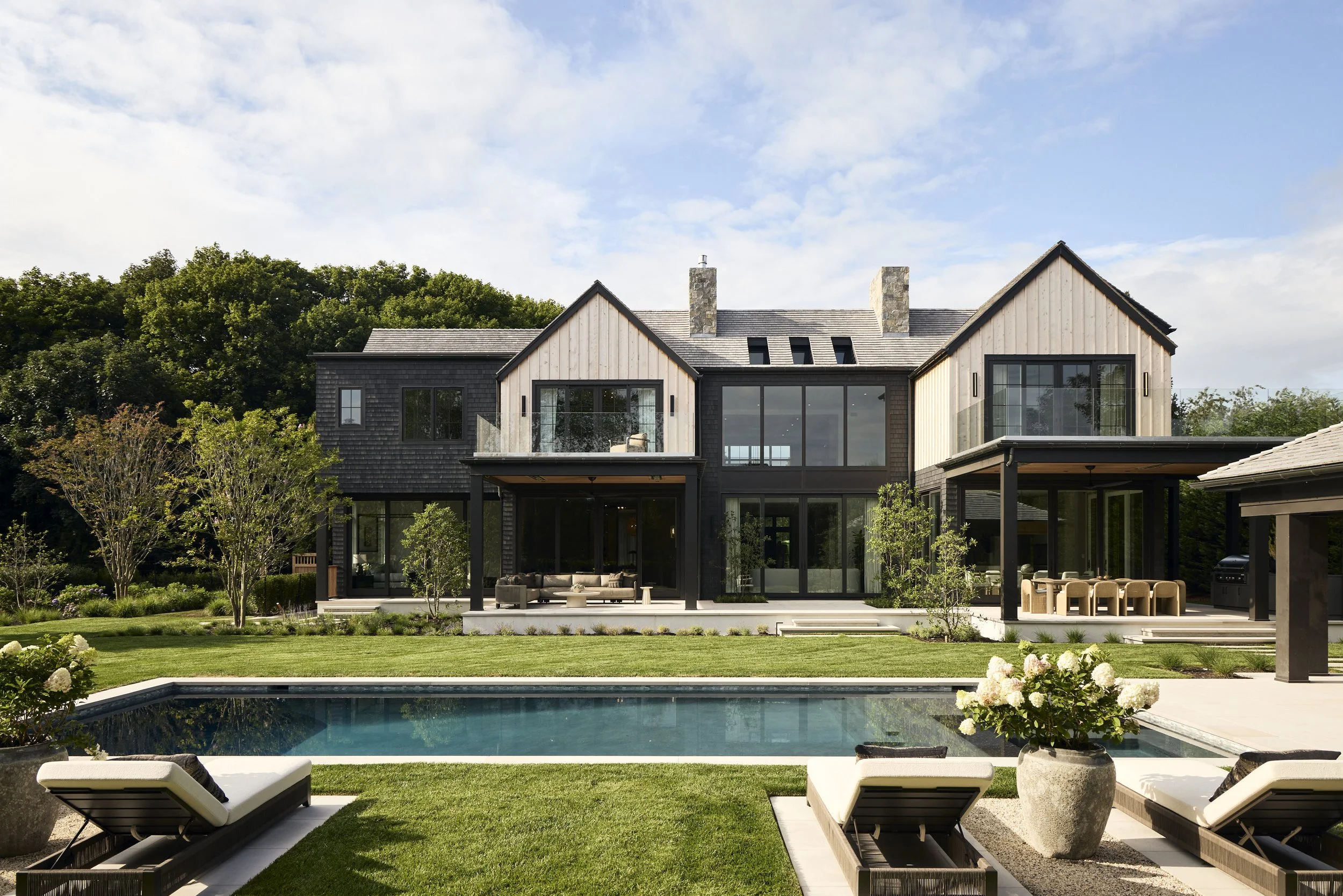 Modern house with a gray roof and large glass windows, surrounded by greenery, pool, and outdoor furniture.