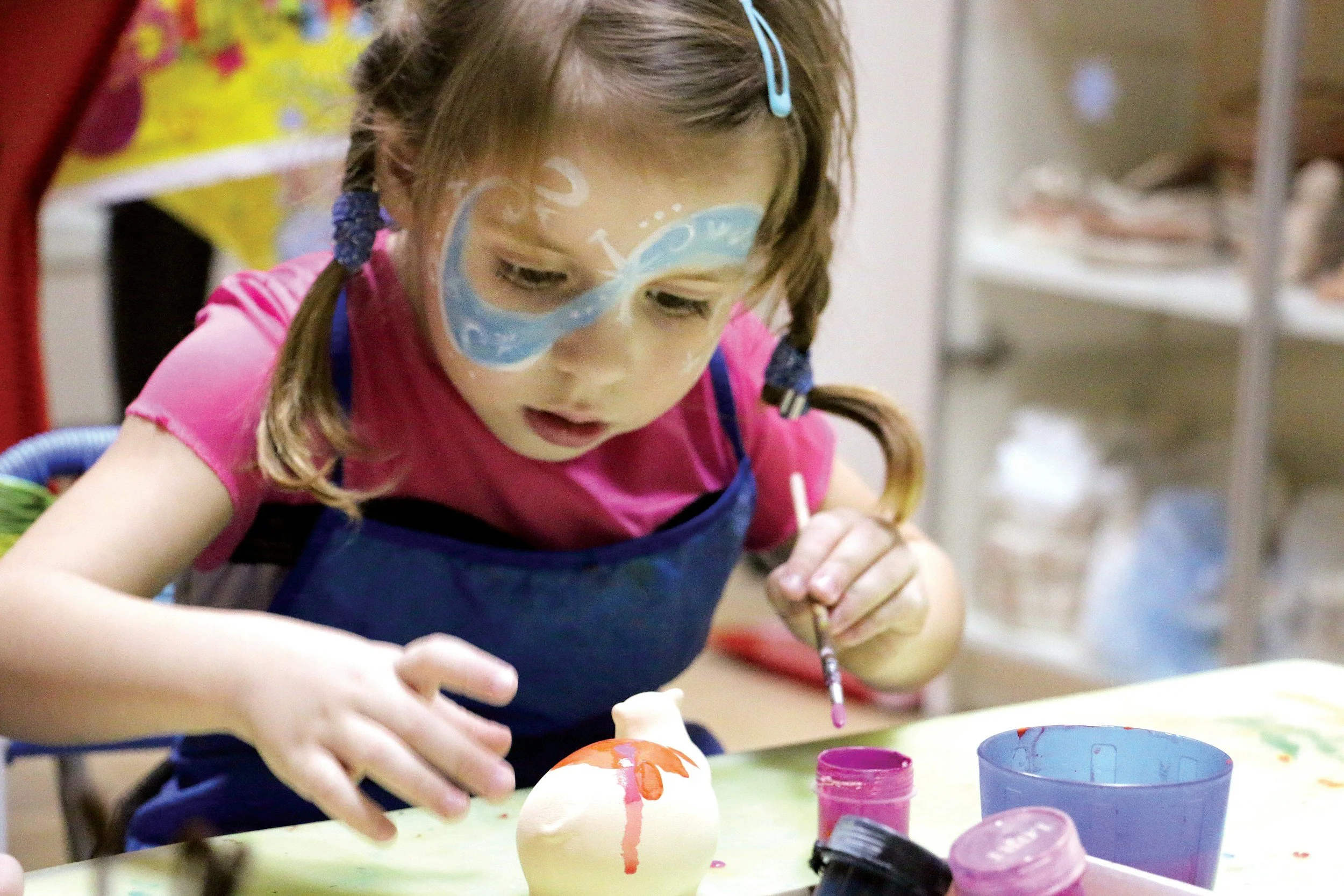A young girl with face paint and braids is painting an egg with orange paint, sitting at a table with containers of paint and art supplies around her.