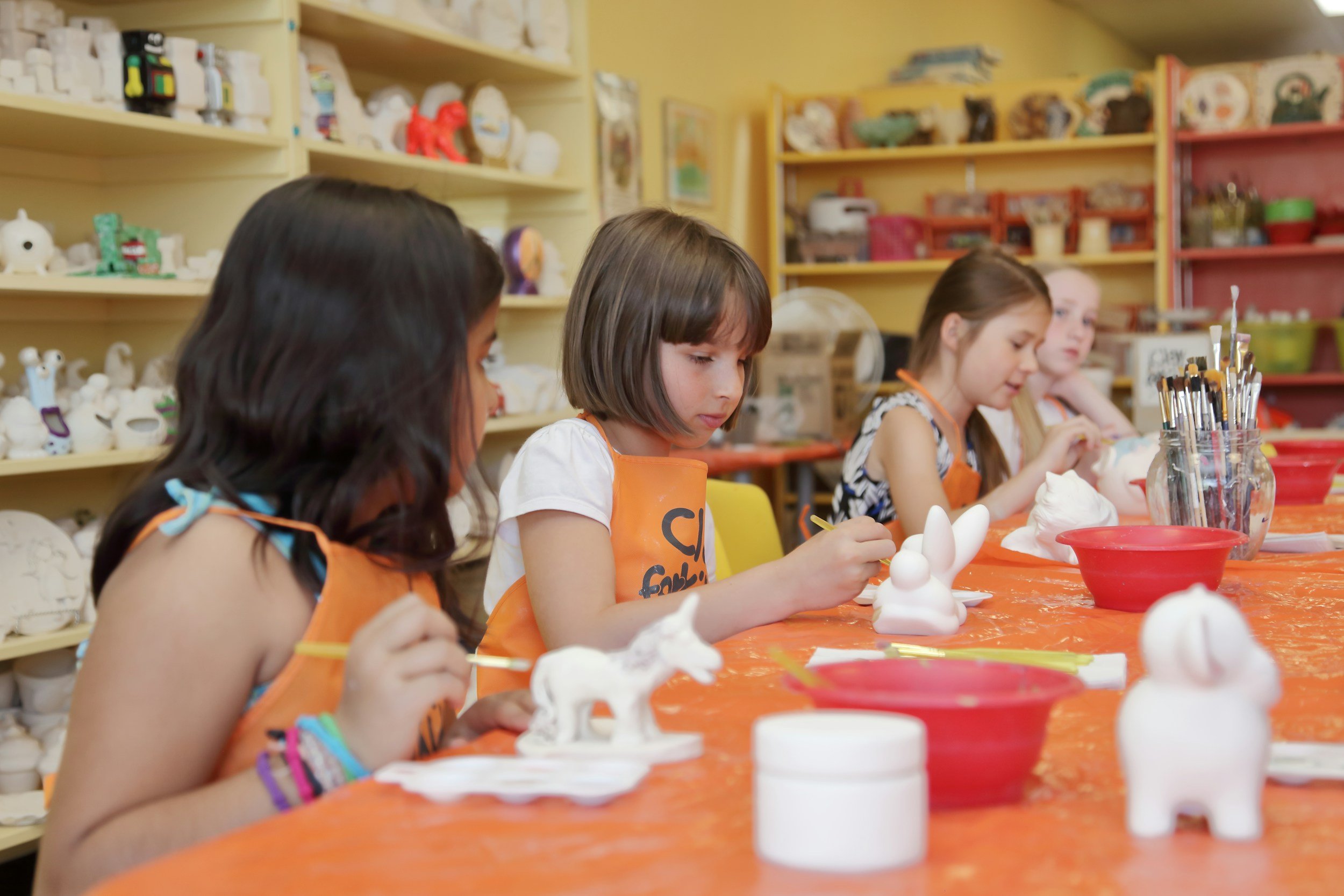Children sitting at a table engaged in painting or sculpting pottery in an art classroom, with shelves of ceramic sculptures and art supplies in the background.