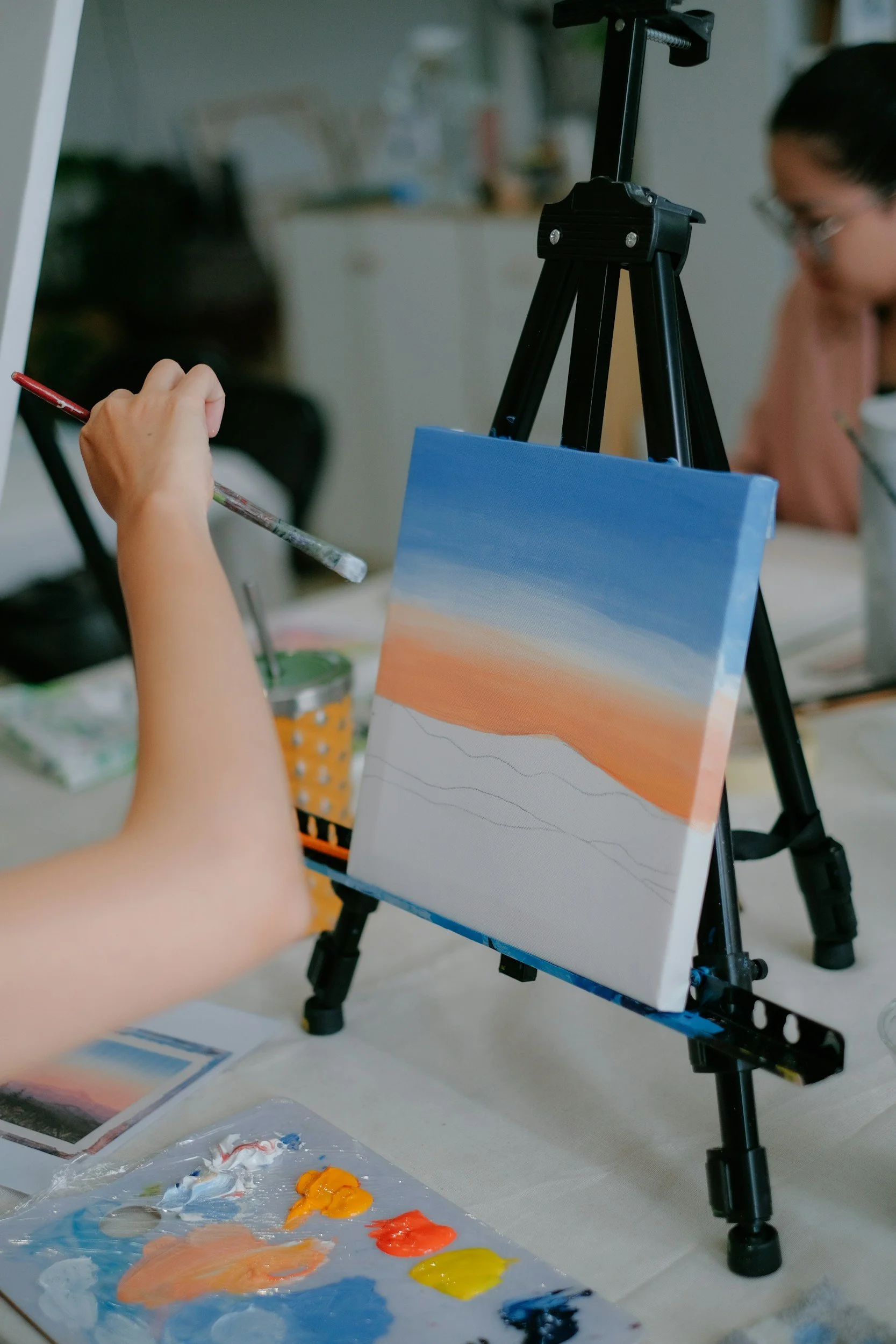 A person painting a landscape scene on a canvas with a gradient sky, using a palette with various colors of paint, while another person watches nearby.