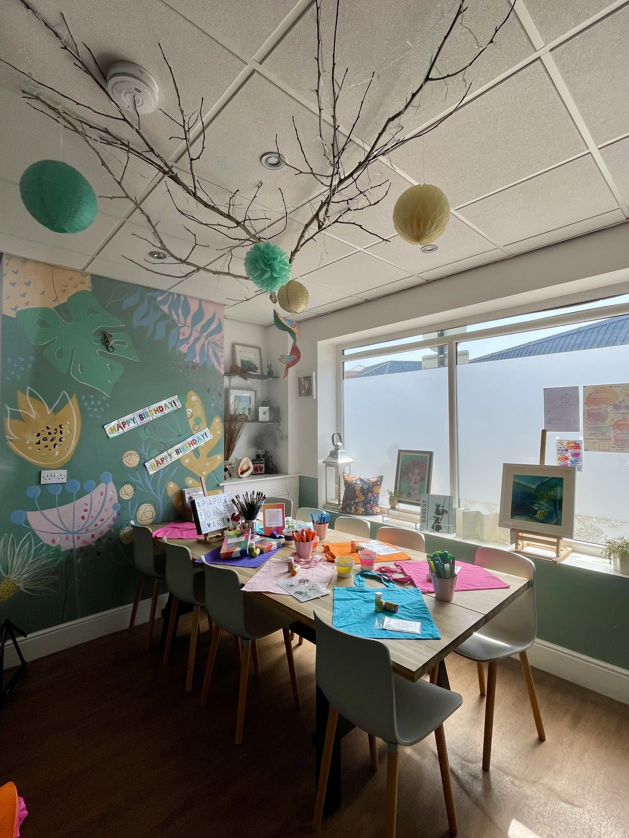 A room decorated for a birthday celebration with a large wooden table covered with colorful craft supplies and decorations. The wall features a mural with large floral and leaf patterns, and there's a adjacent window allowing natural light into the room.