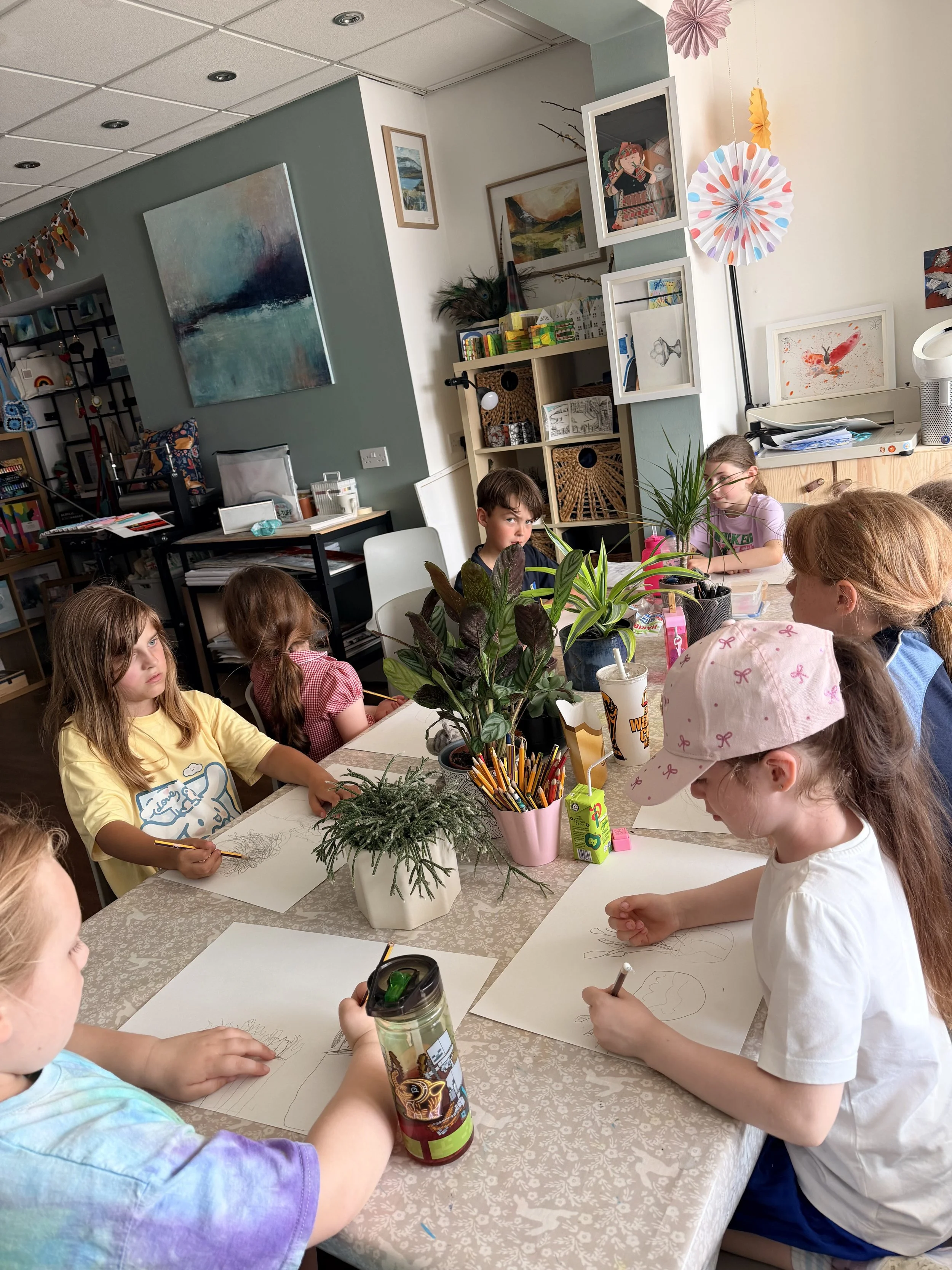 Children sitting around a table, drawing on white sheets of paper, with a potted plant in the center of the table. The classroom wall has various artworks and decorations, including framed pictures, a lamp, and colorful paper fans.