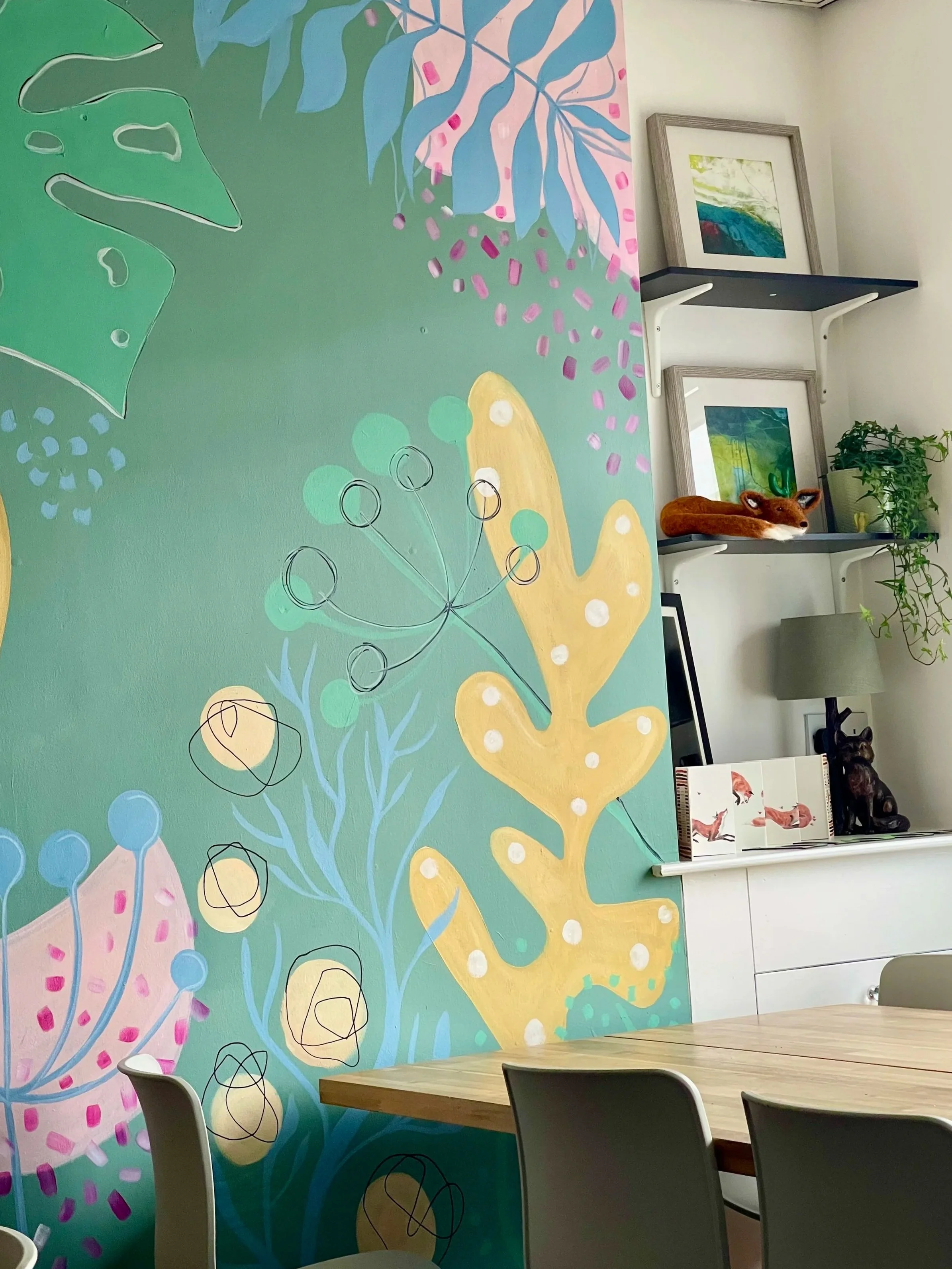 A colorful painted wall with abstract floral and leaf designs in pastel colors like pink, blue, yellow, and green, next to a white wall with framed artwork, shelves, and decorative items including a plush fox and a potted plant in a modern indoor setting.