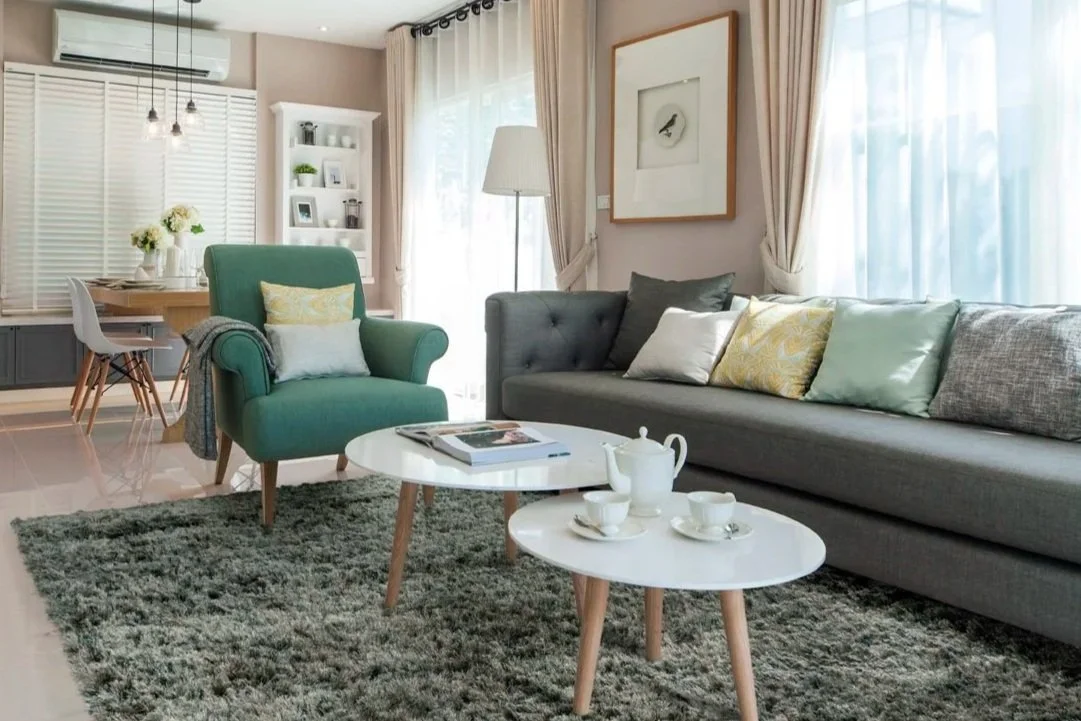 Een woonkamer met een lange grijze bank, een groene armstoel, een hoeklamp, poesjes voor de muur, een wit boekenkast, een eettafel met bloemen, en een glazen deur met gordijnen.