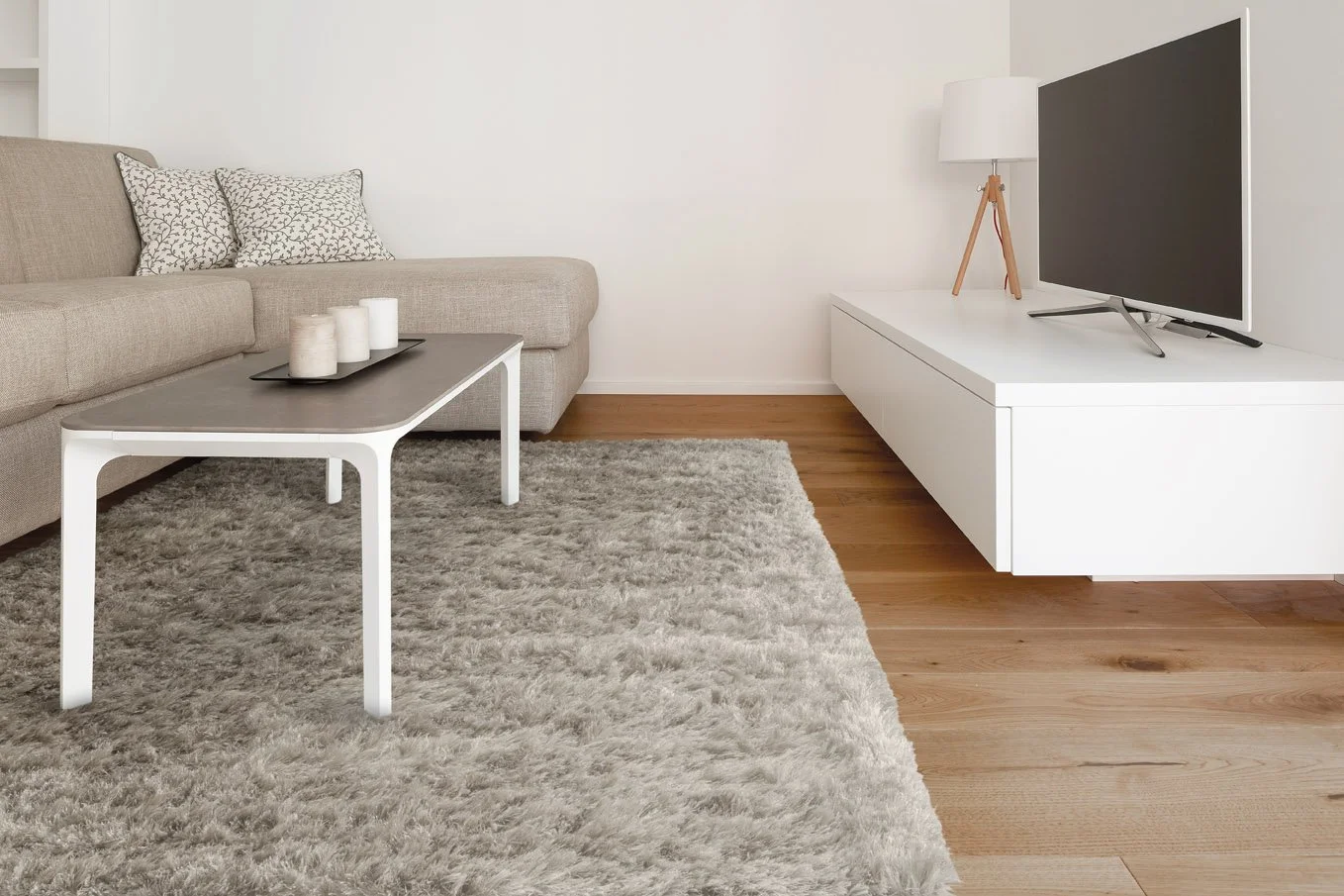 Woonkamer met beige bank, wit tv-meubel met tv, houten vloer, grijze tapijt, bloemenkussen en een salontafel met kaarsen.