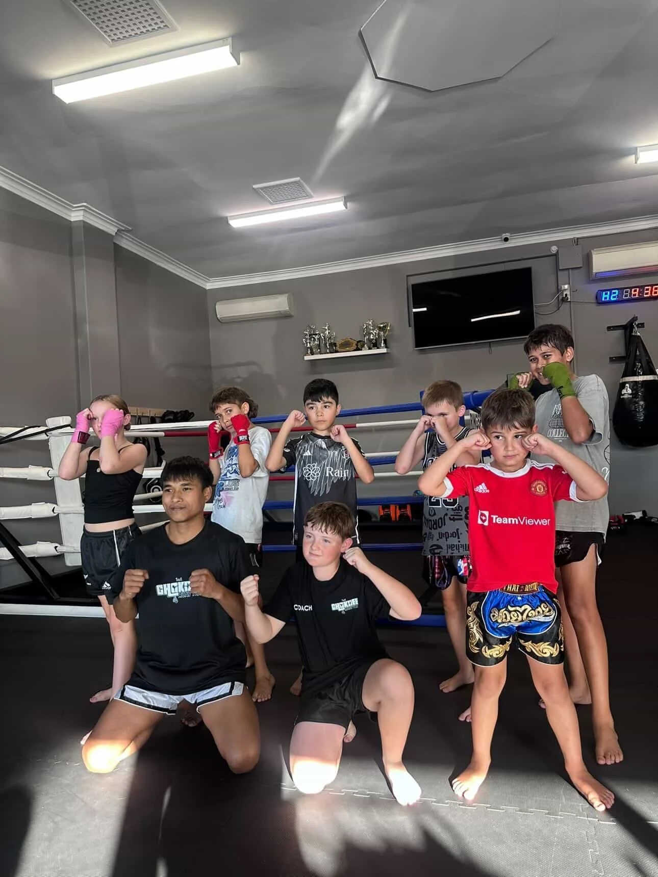 A group of young children and teenagers in boxing poses inside a gym, with some wearing boxing gloves and shorts, in front of boxing rings and trophies.